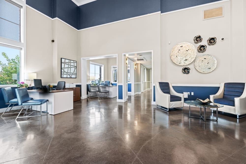 Modern office lobby interior with high ceilings, navy and neutral-toned walls, polished concrete floors, and contemporary decor. The space includes a reception desk with floral arrangements, blue and chrome guest chairs, abstract wall art pieces, and natural light streaming in through large windows. An adjoining hallway with matching decor is visible in the background at Marq Inverness in Englewood, CO.