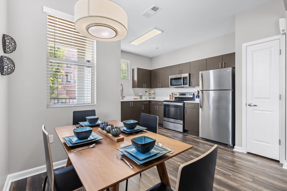 Modern kitchen and dining area with natural light coming through the window, featuring stainless steel appliances, wooden cabinets, a dining table set for four with blue dishes, and decorative black wall hangings at Marq Inverness in Englewood, CO.