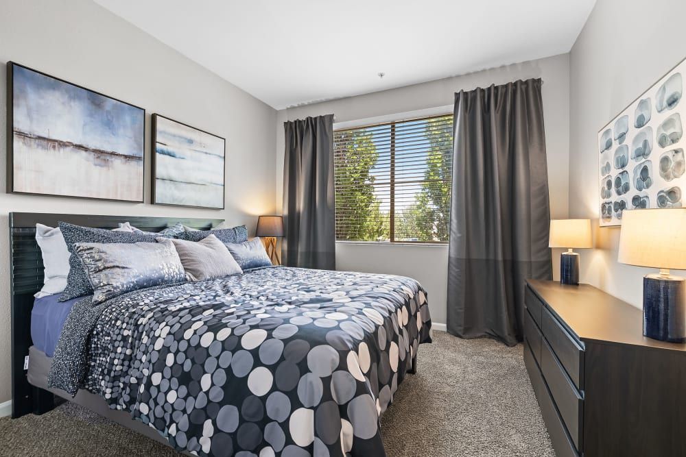 A modern bedroom with gray walls and coordinated dark gray curtains. The room features a bed with a patterned comforter and multiple pillows, flanked by nightstands with navy blue lamps. Above the bed are two framed abstract paintings, and a third artwork is positioned on the adjacent wall by the window that lets in natural light at Marq Inverness in Englewood, CO.