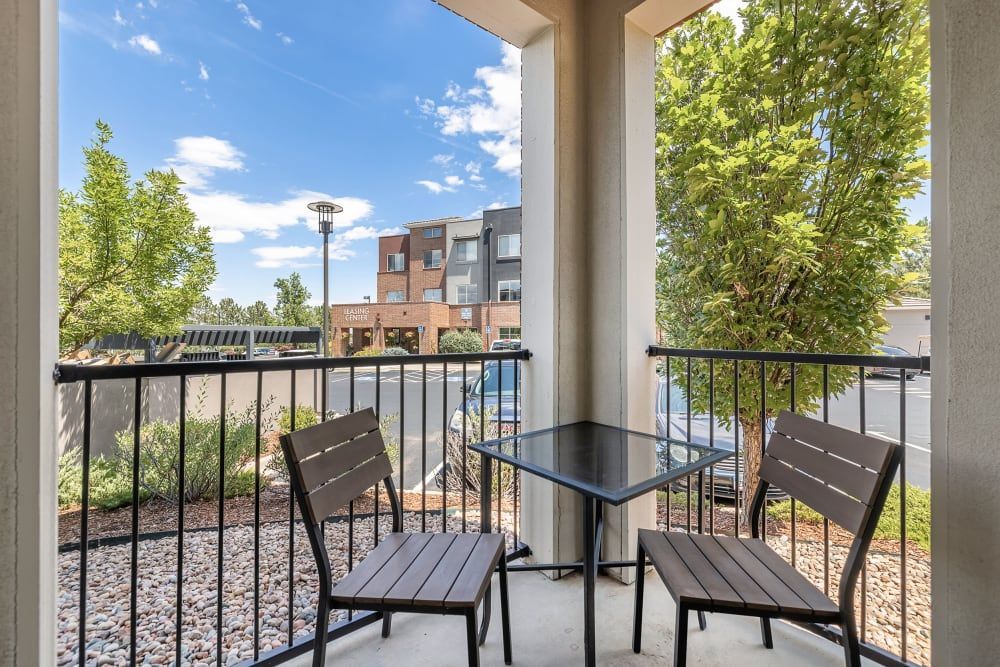 View from a cozy apartment balcony with a small patio table and two chairs overlooking a landscaped area with trees, a parking lot, and a building with a 'Leasing' sign in the distance on a sunny day at Marq Inverness in Englewood, CO.