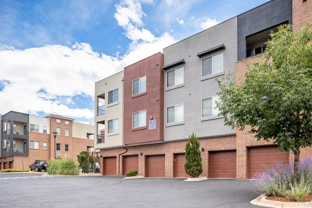 Modern multi-story residential buildings with a mix of brown and grey facades, featuring individual garages on the ground floor. A parking lot is in the foreground, with lush green trees and decorative shrubs along the edges. The sky is partly cloudy at Marq Inverness in Englewood, CO.