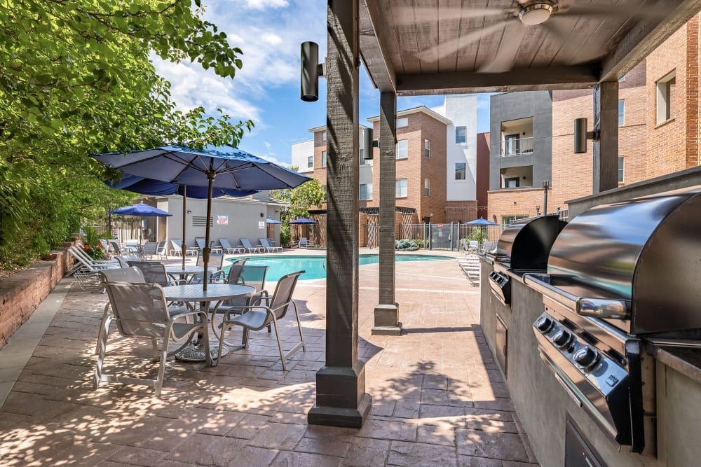 Outdoor residential communal area with a swimming pool, lounge chairs under blue umbrellas, and a cooking area with a large barbecue grill. The space is surrounded by multi-story brick apartment buildings, with lush green foliage to the left and clear blue skies above at Marq Inverness in Englewood, CO.