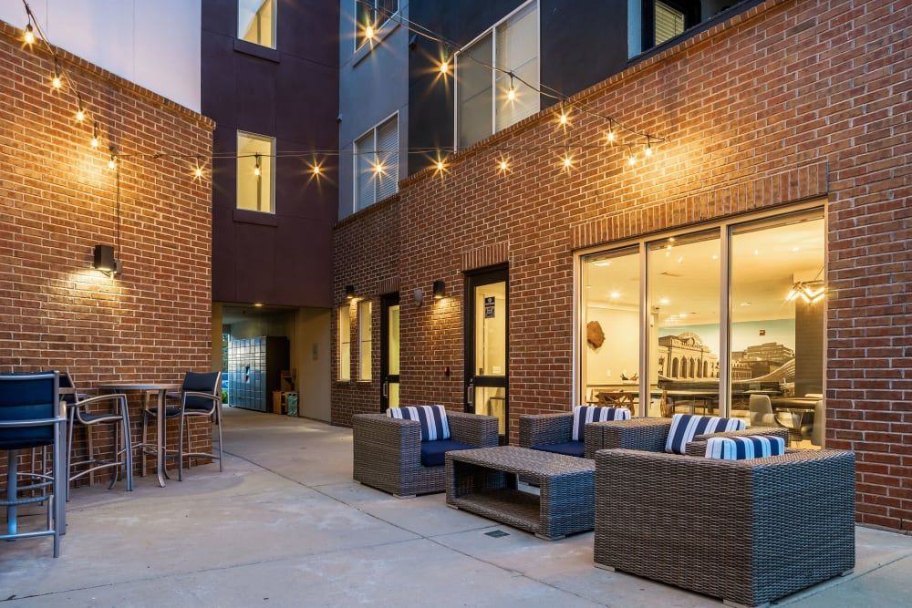 A cozy outdoor seating area with modern wicker furniture, including sofas with blue striped cushions and a high table with bar stools. The setting is flanked by red brick walls of adjacent buildings, with warm string lights overhead, and large windows looking into a well-lit interior space at Marq Inverness in Englewood, CO.