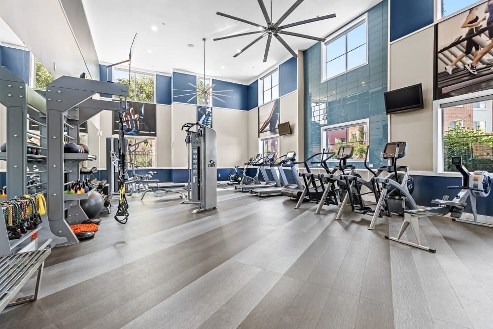 Modern gym interior with a variety of equipment including treadmills, elliptical machines, and a multi-station weight machine. The room features high ceilings with a large ceiling fan, blue and white walls, and floor-to-ceiling windows that let in natural light. Television screens are mounted on the walls, and a rack with exercise balls and free weights is visible on the left at Marq Inverness in Englewood, CO.