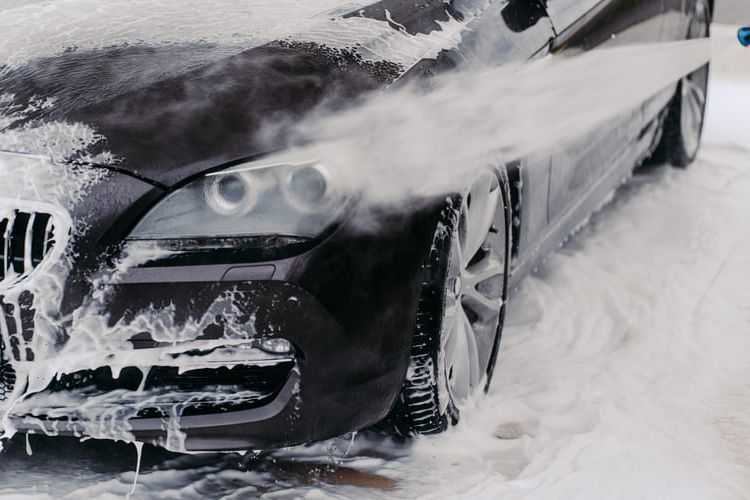 A person is snow foam washing a black car with a high pressure washer.