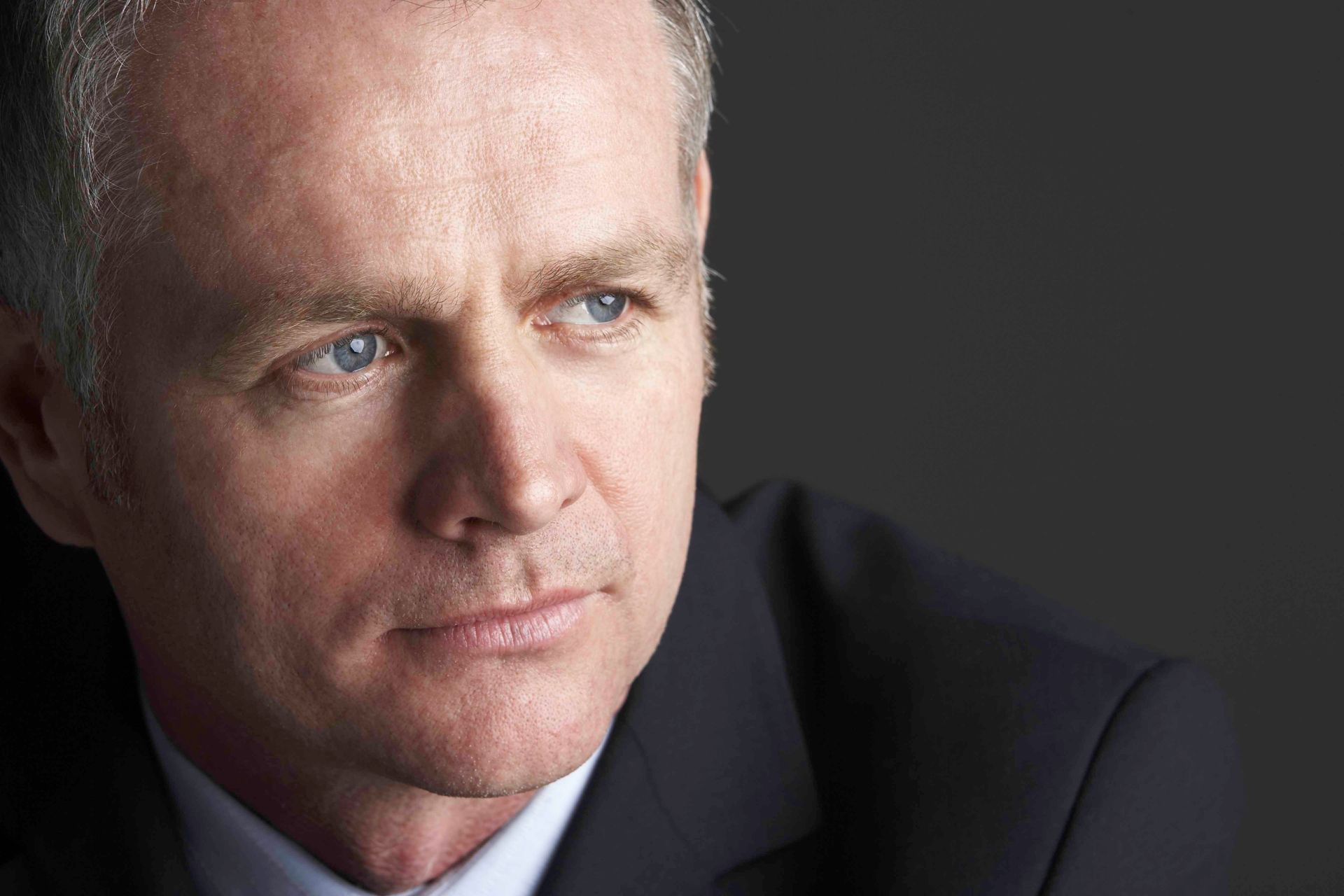 Man in a suit looks off-camera with a thoughtful expression; against a dark background.