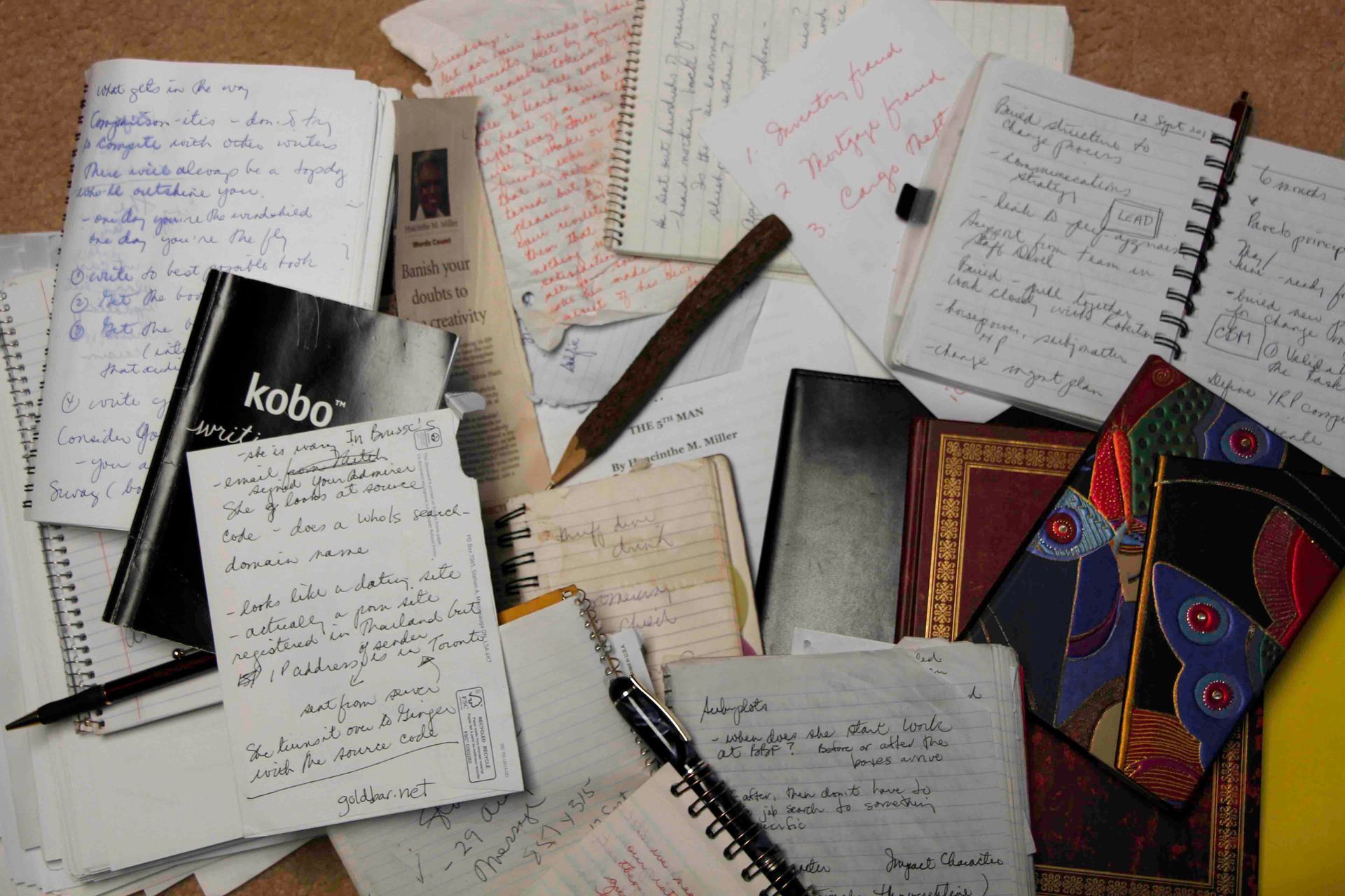 Pile of open notebooks and papers with handwritten notes; a pen, and a Kobo e-reader are visible.