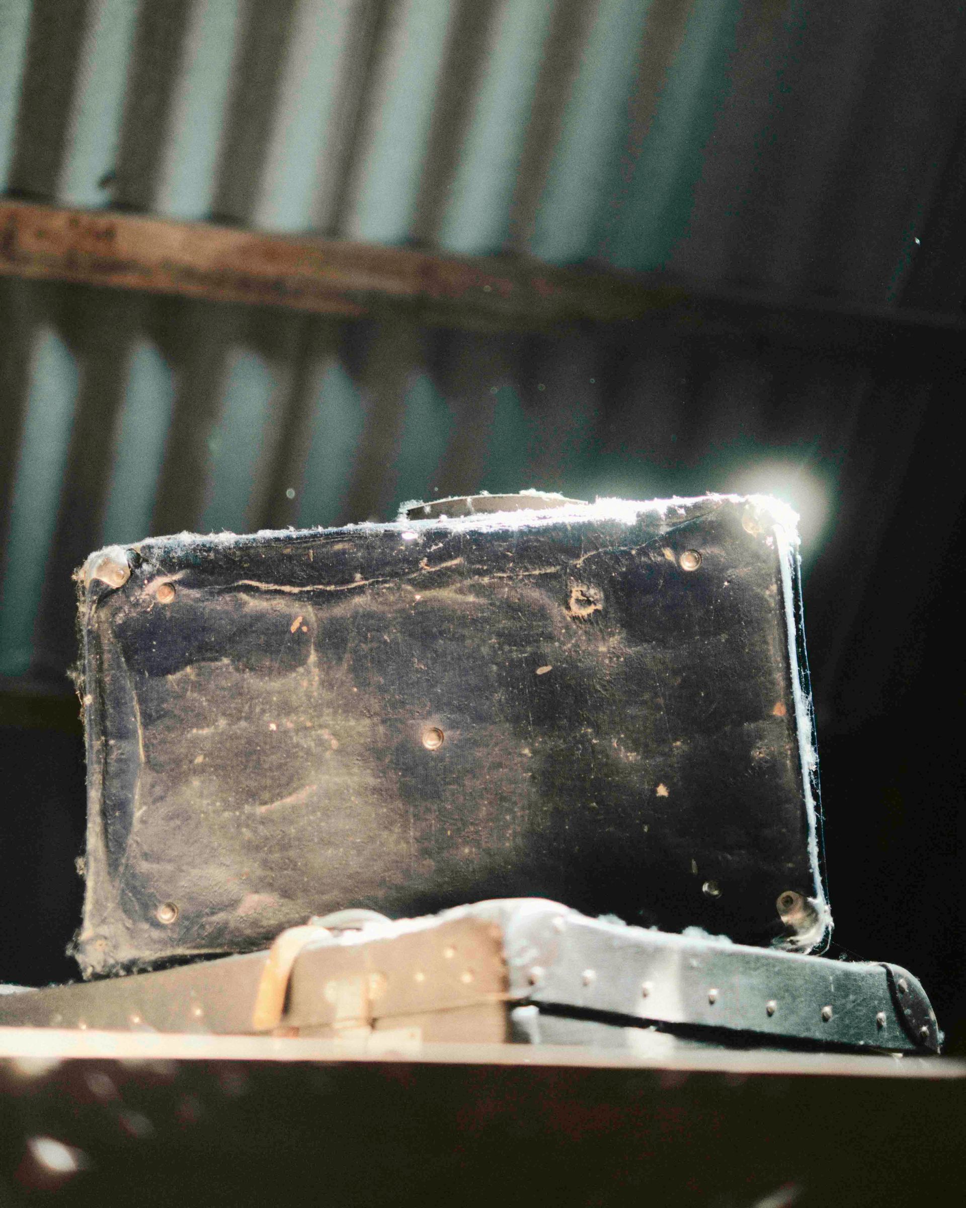 Old, dusty black briefcase on a shelf under a corrugated metal roof. Sunlight shining.