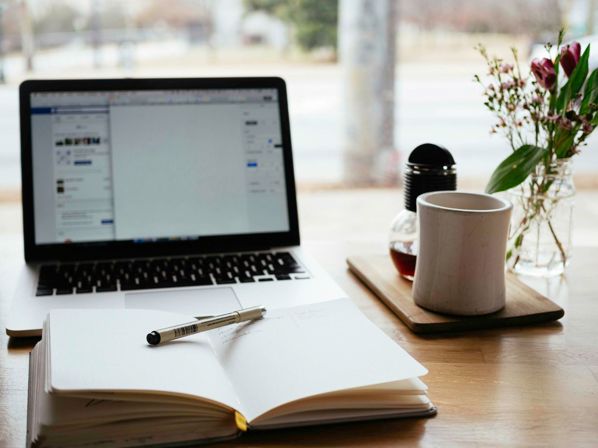 Laptop, open notebook, coffee cup, flowers, and pen on a wooden table near a window.