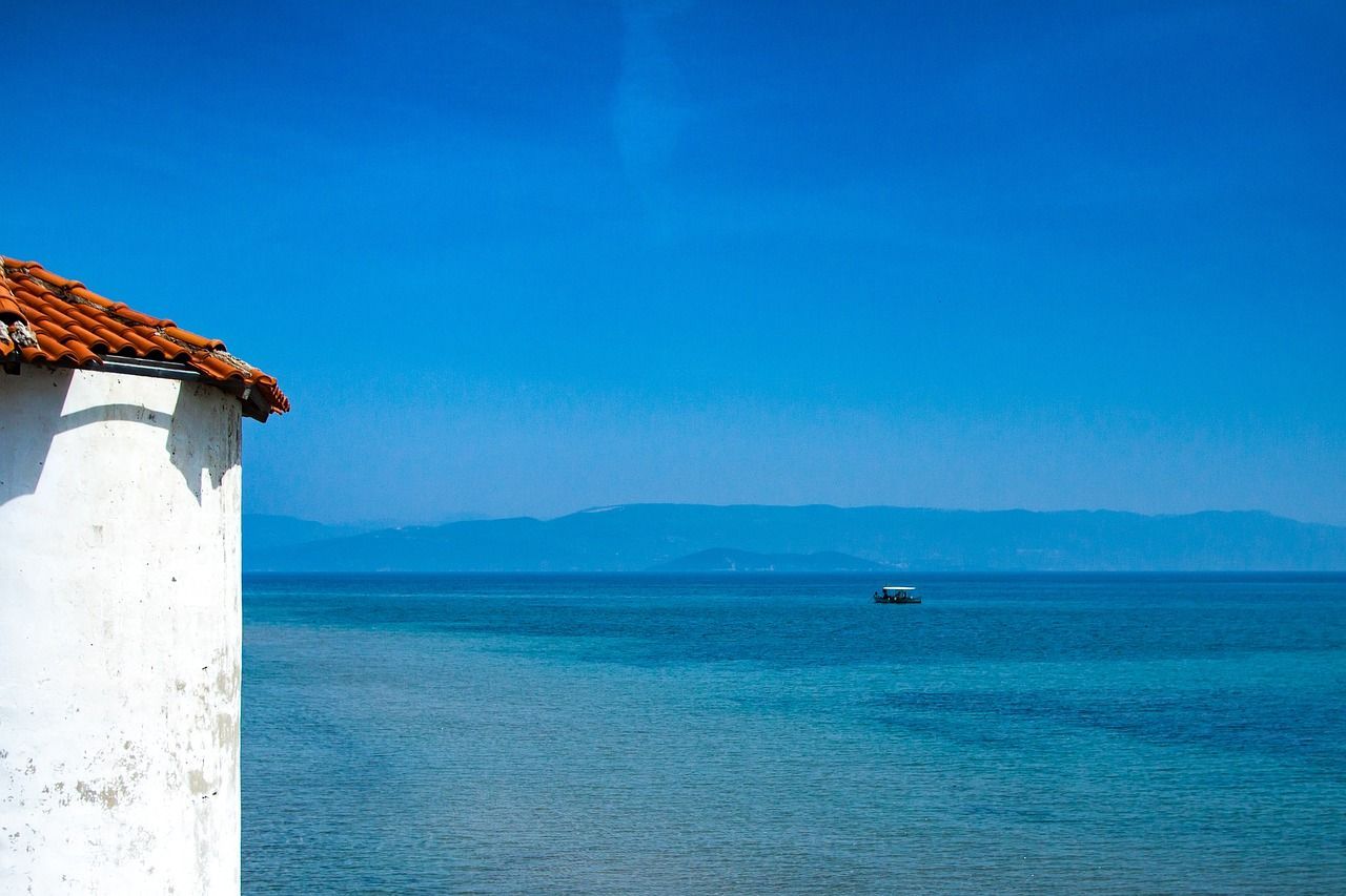 White building corner with orange roof by blue sea, small boat on water, mountains in the distance.