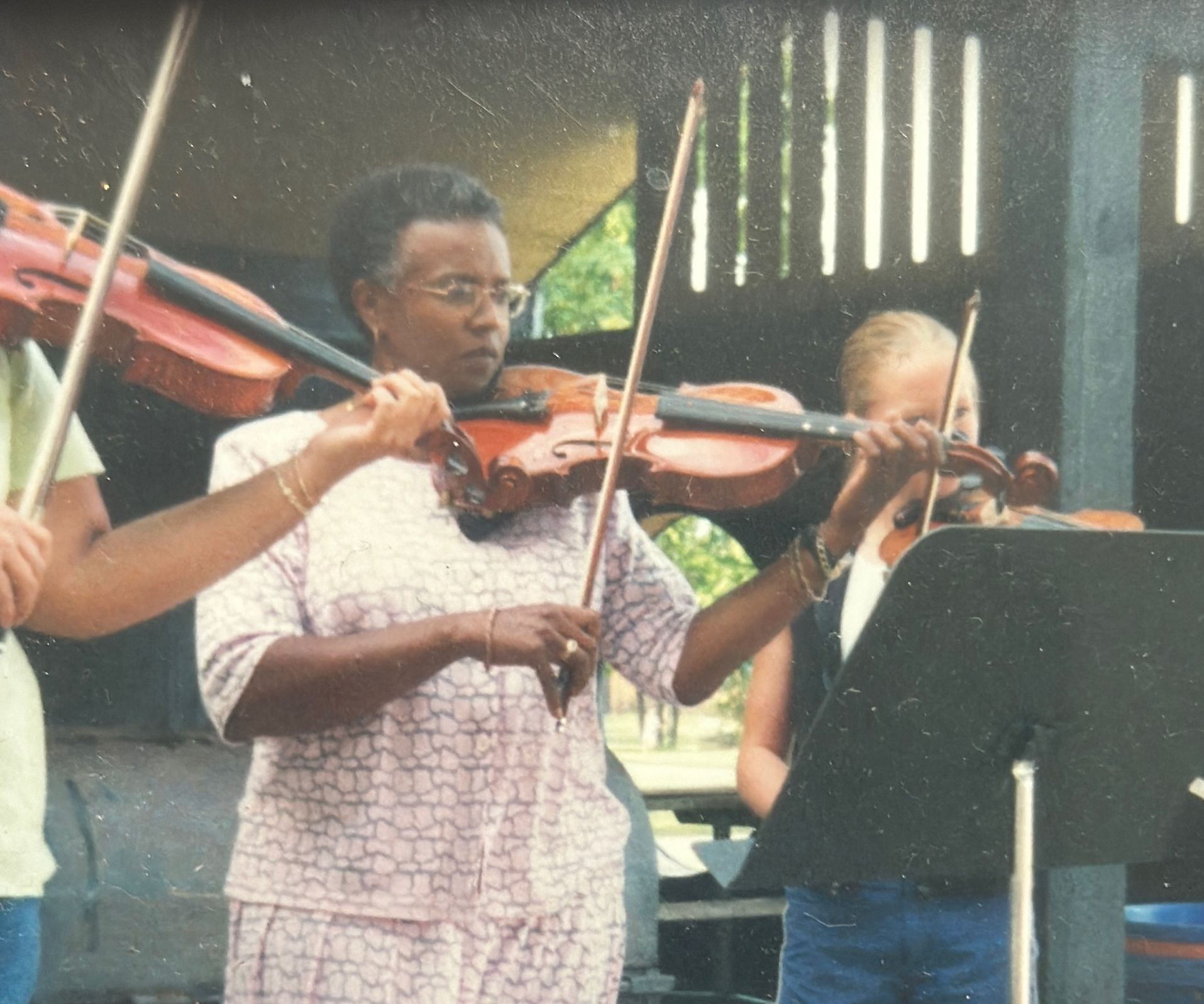 Woman playing the violin outdoors with other musicians; wearing a pink patterned suit.