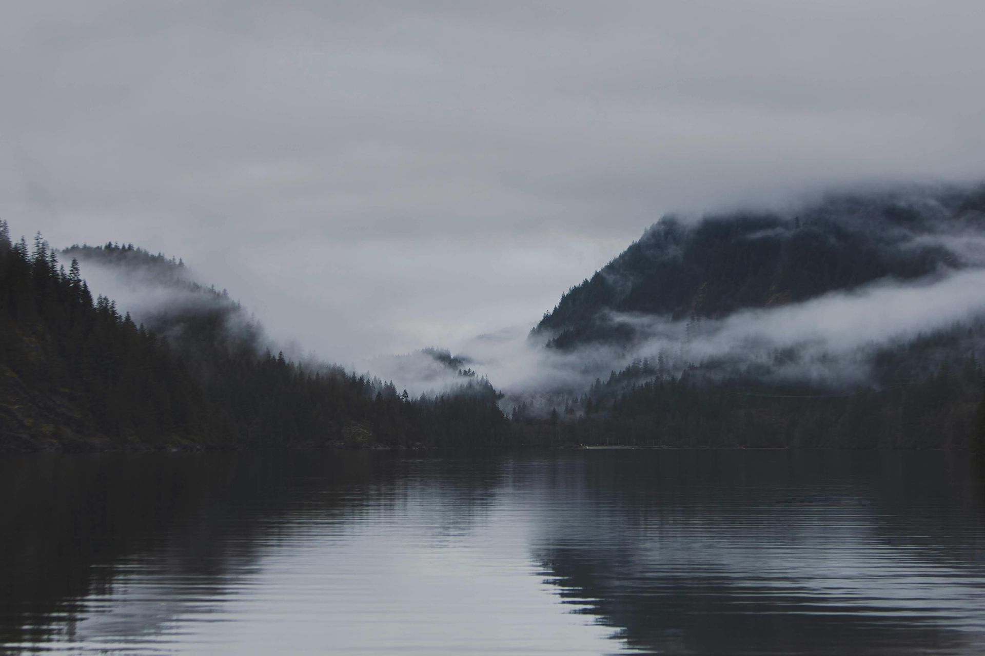Gray lake reflects mist-covered mountains, shrouded in clouds.