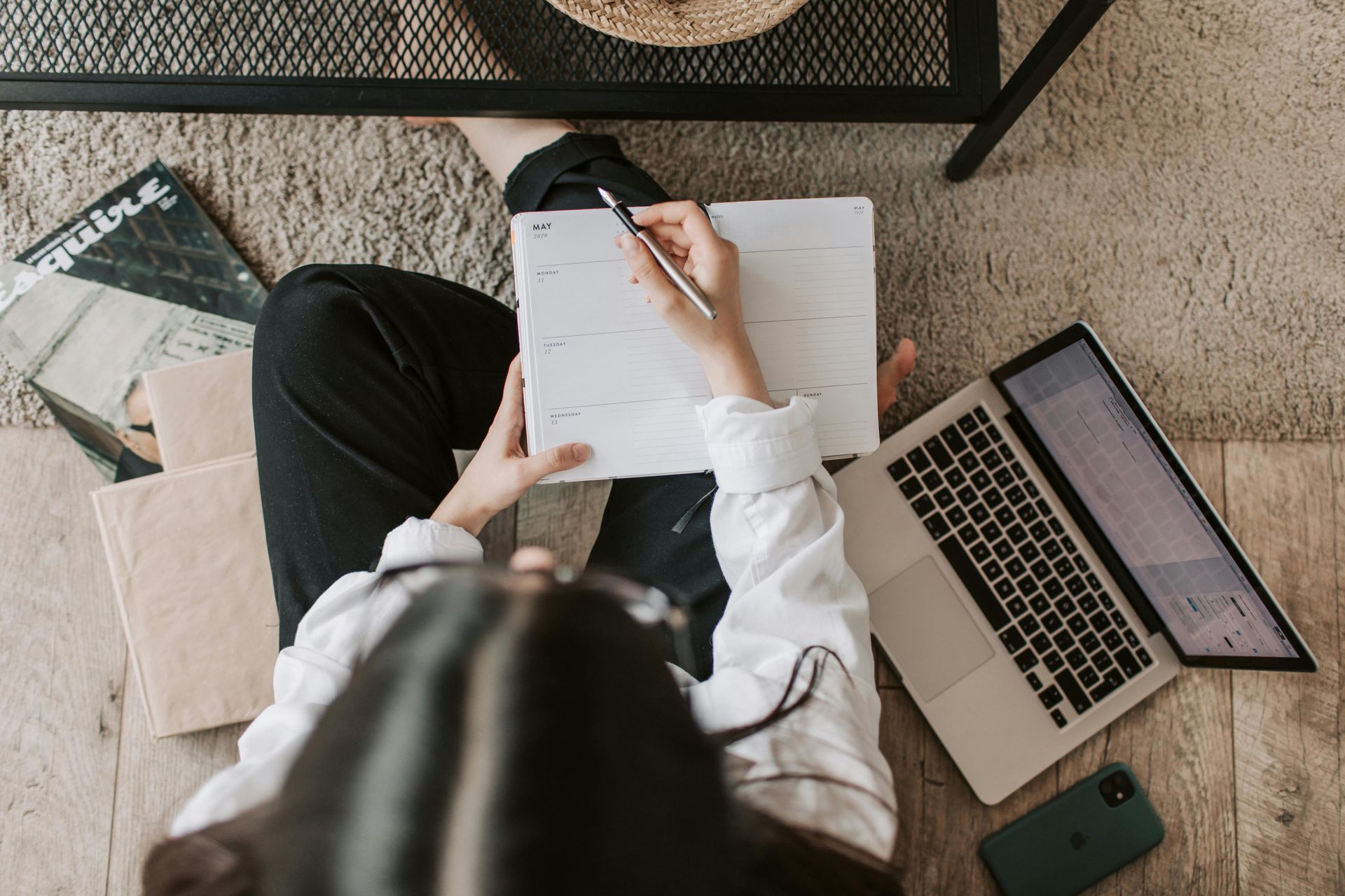 Person seated on floor, writing in planner next to laptop and phone; books nearby.