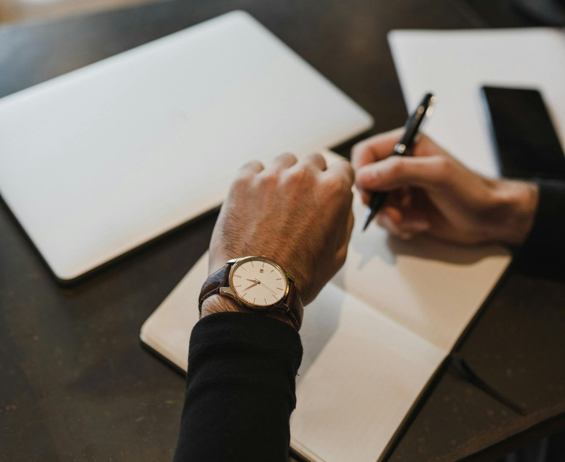 Person writing in a notebook, checking watch. Laptop and phone on the table.