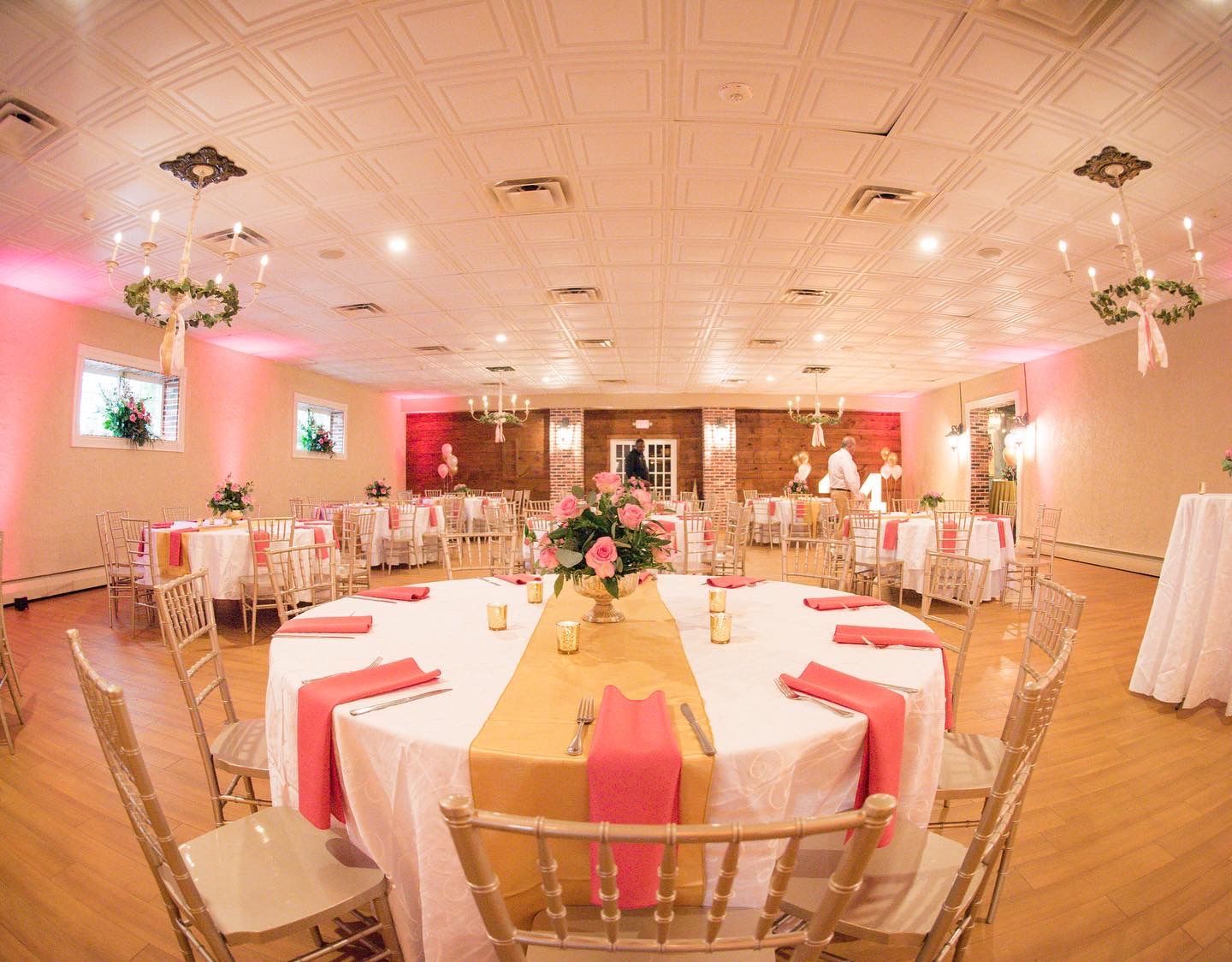 A well-lit reception hall with round tables set for a celebration. Pink and gold decor.