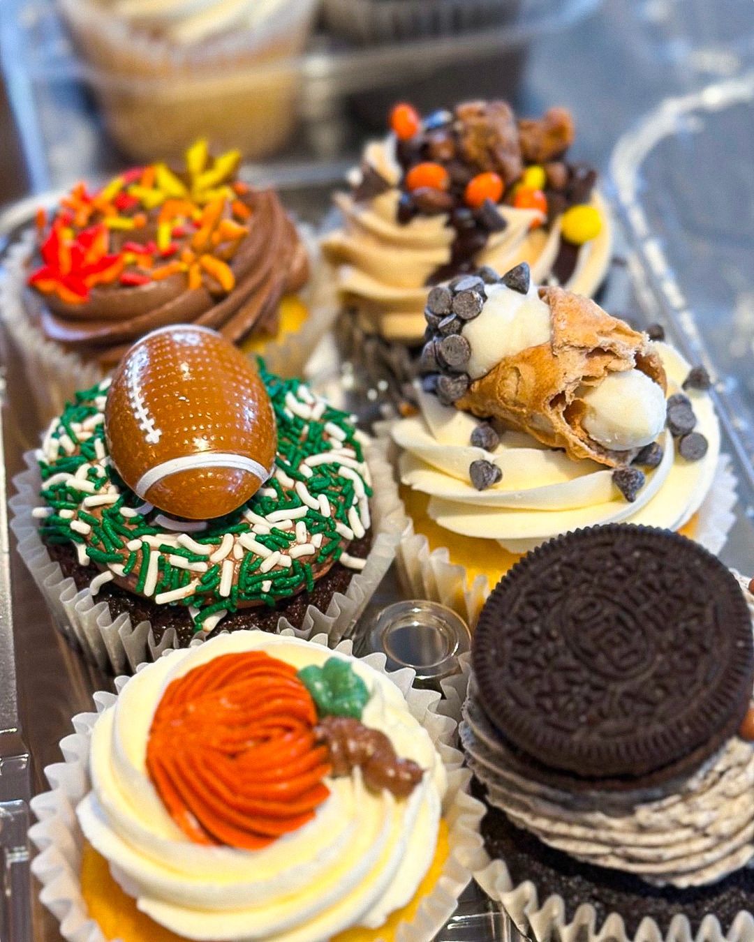 Cupcakes in a container: various flavors with decorative toppings, including football, cannoli, and fall leaves.