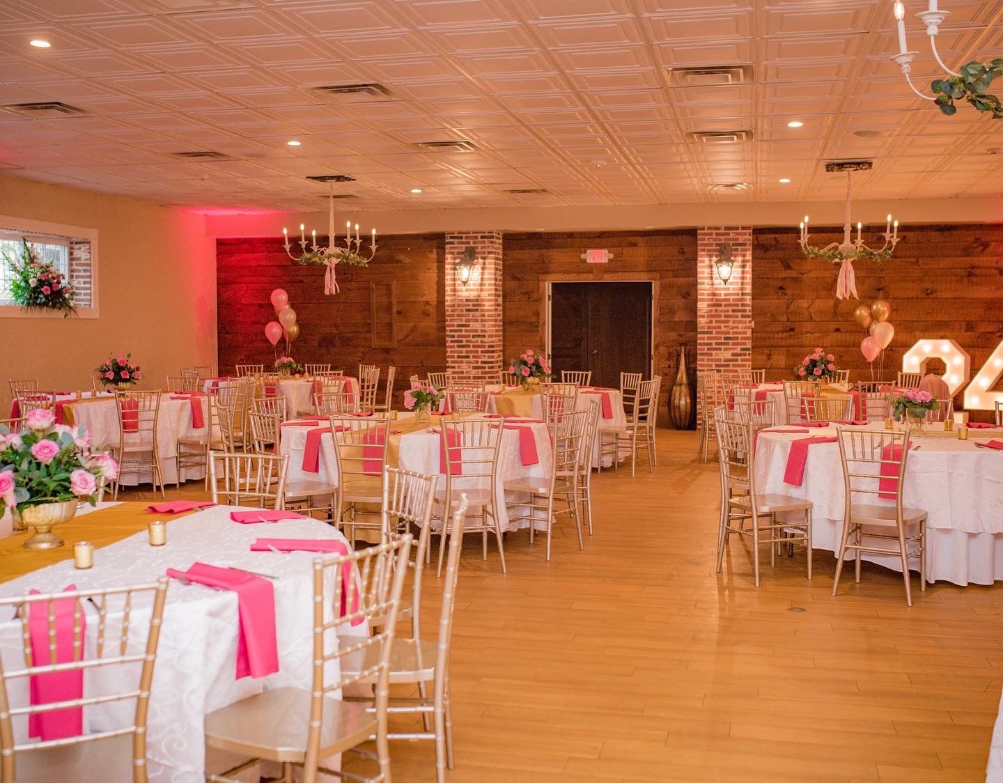 Banquet hall set for an event; round tables with pink and gold decor, wood floors, and chandeliers.