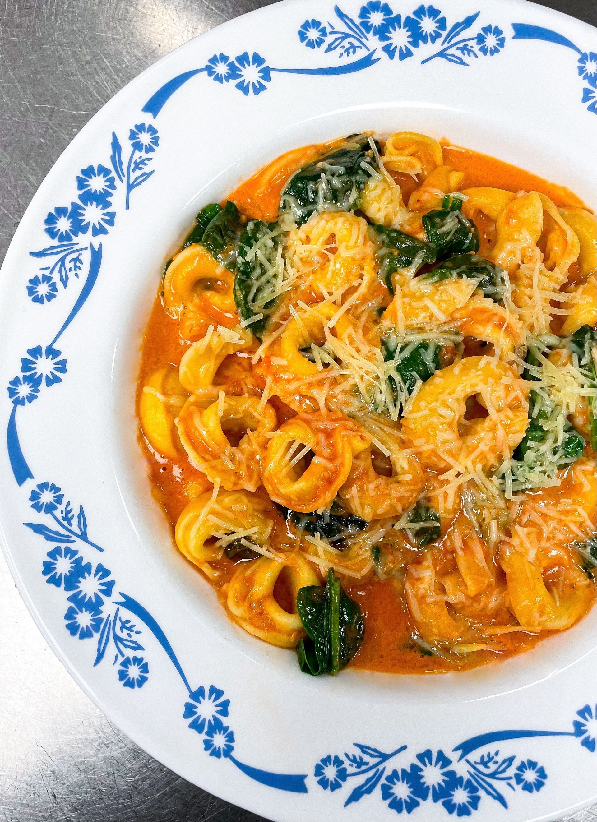 Pasta dish in white bowl with blue floral design, orange sauce, green spinach, and grated cheese.