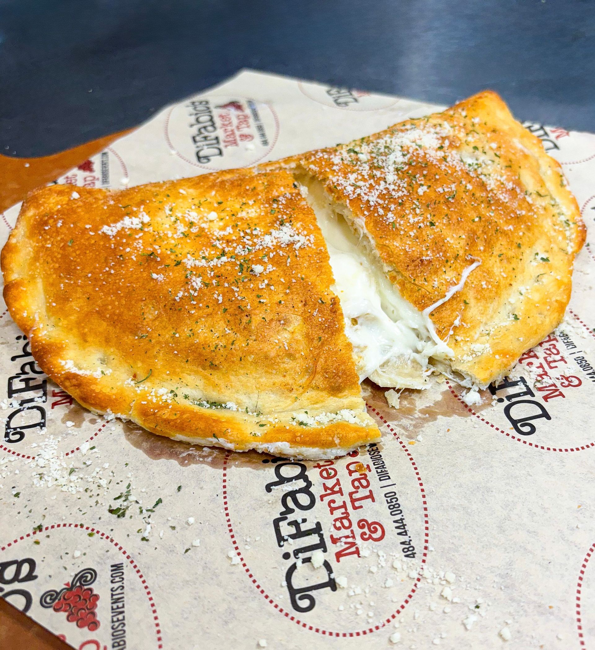 Golden-brown calzone cut open, revealing melted cheese inside. Sprinkled with herbs, on printed paper.