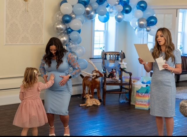 Baby shower: Pregnant woman in blue dress, child touches belly, woman reads, balloons.