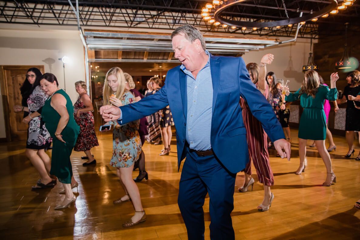 Man in blue suit dances on a wooden floor, surrounded by other people in a lit room.