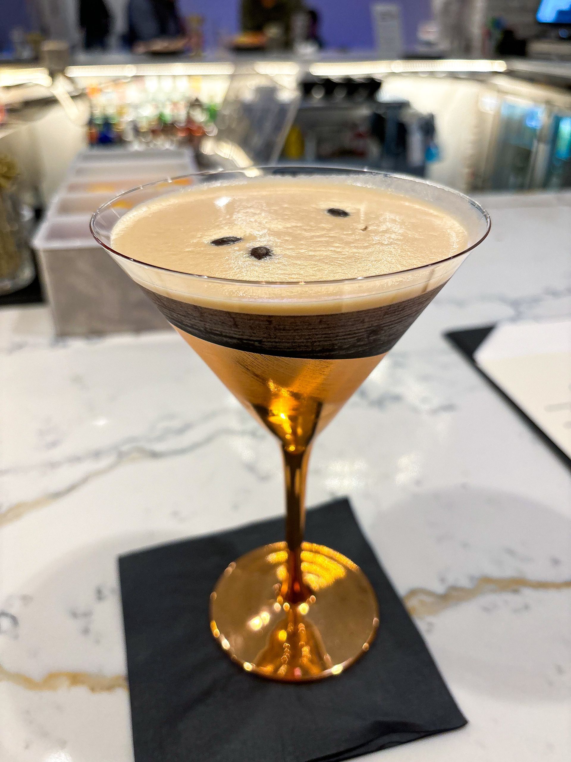 Espresso martini in a gold glass on a bar, garnished with coffee beans, sitting on a black coaster.