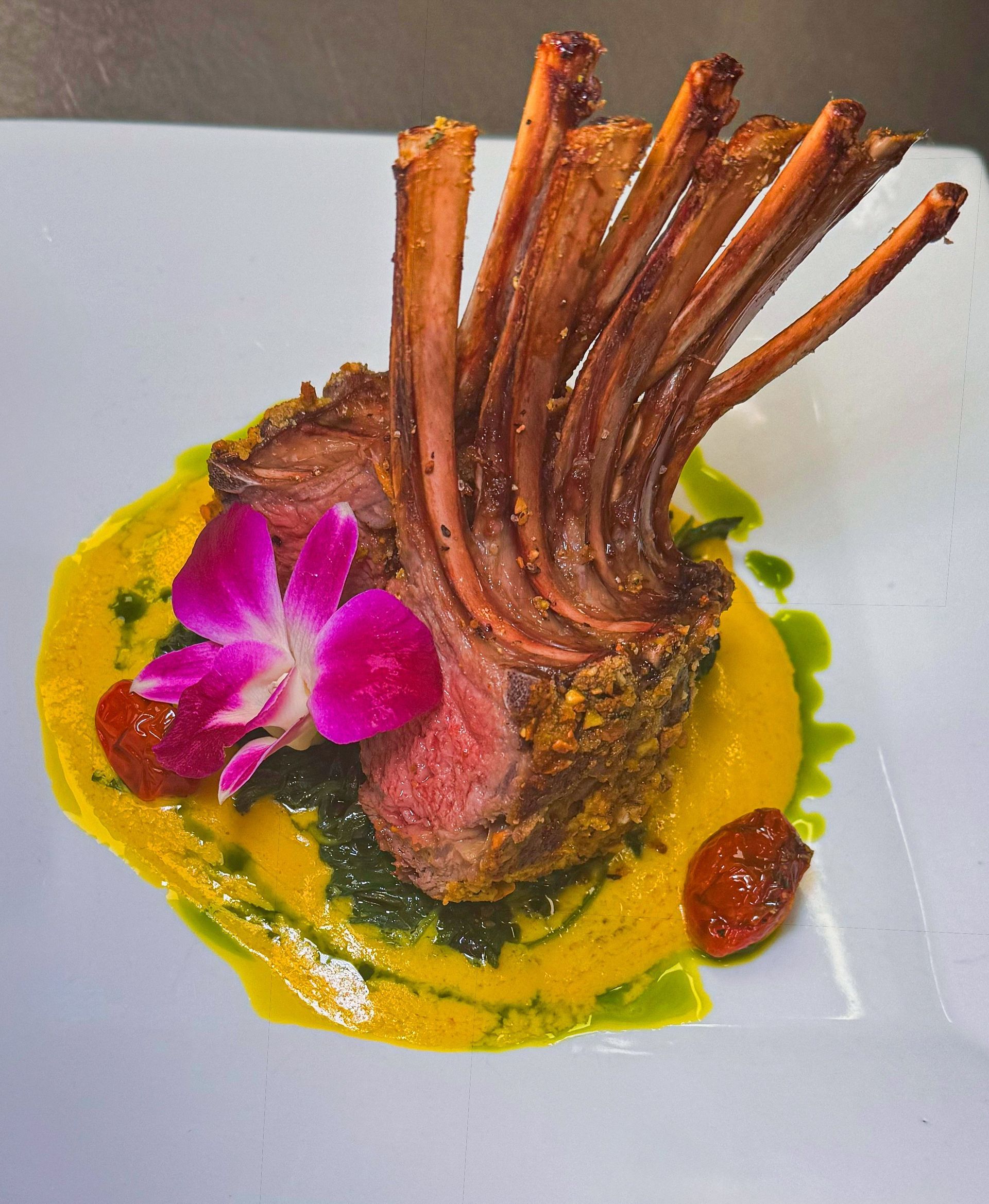 Rack of lamb with a yellow sauce, pesto, cherry tomatoes, and a flower on a white plate.