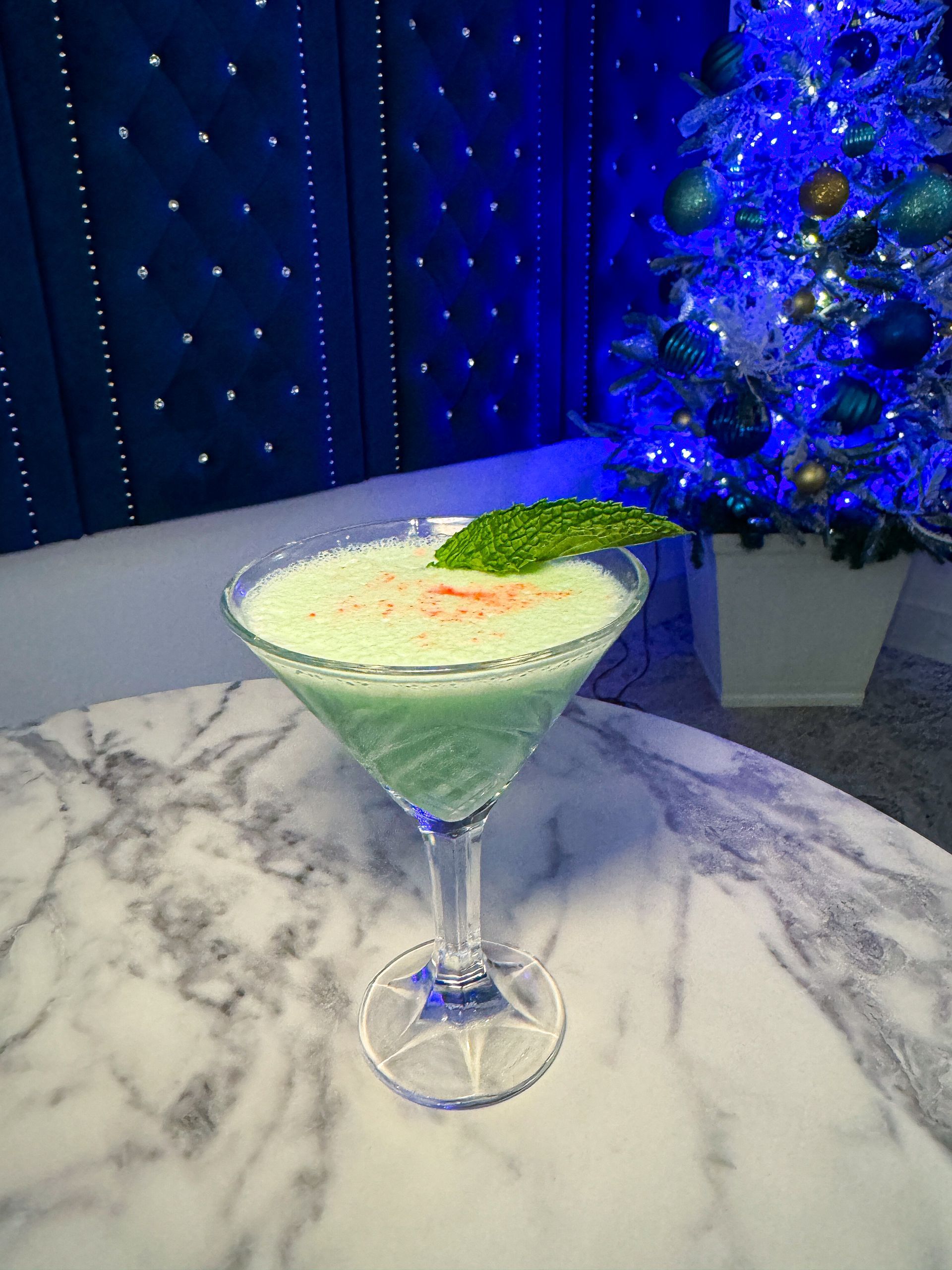 Green cocktail in a martini glass, garnished with mint and paprika, on a marble table with a decorated Christmas tree in the background.