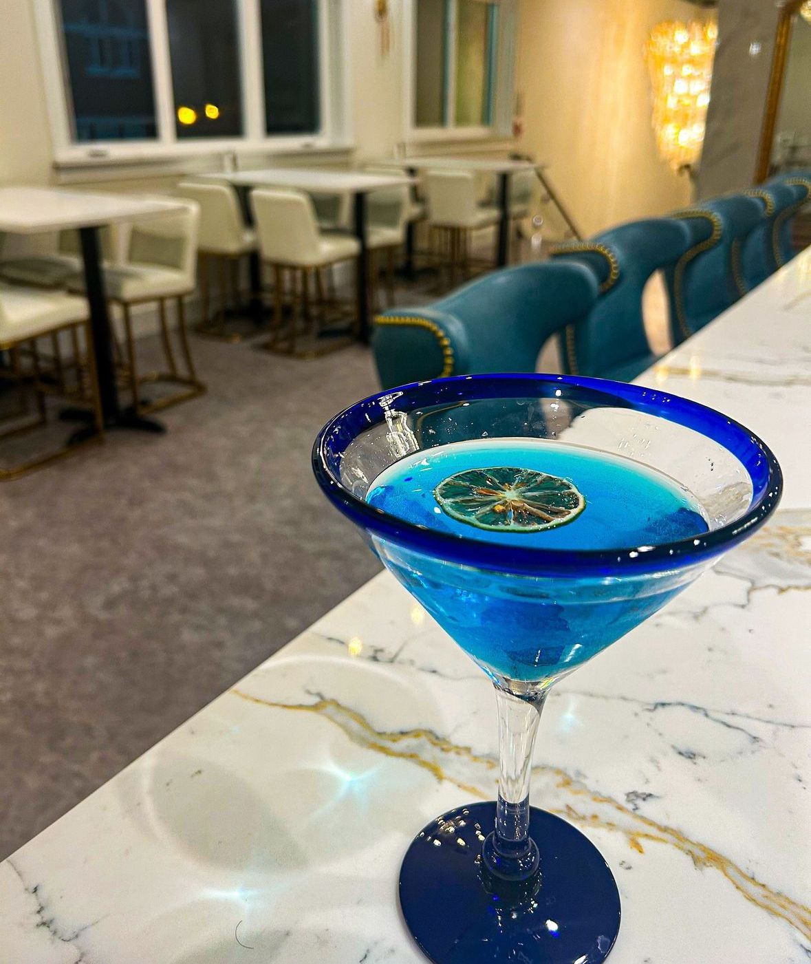 Blue cocktail in a martini glass with a blue rim, set on a white marble bar. A restaurant is in the background.