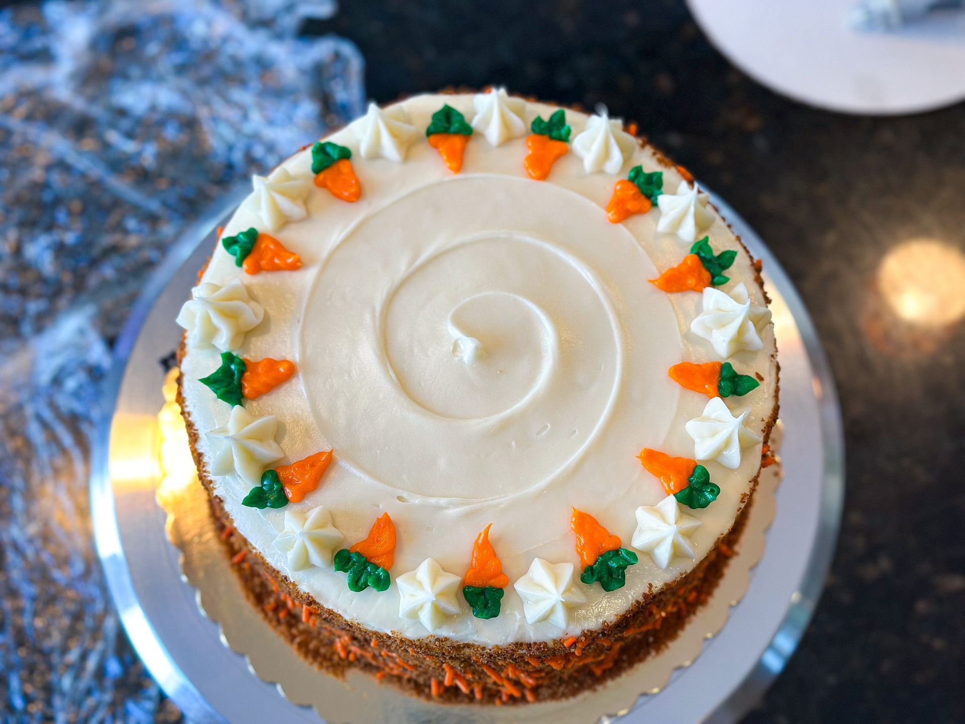 Carrot cake with white frosting, decorated with orange carrot and green leaf frosting details, on silver platter.
