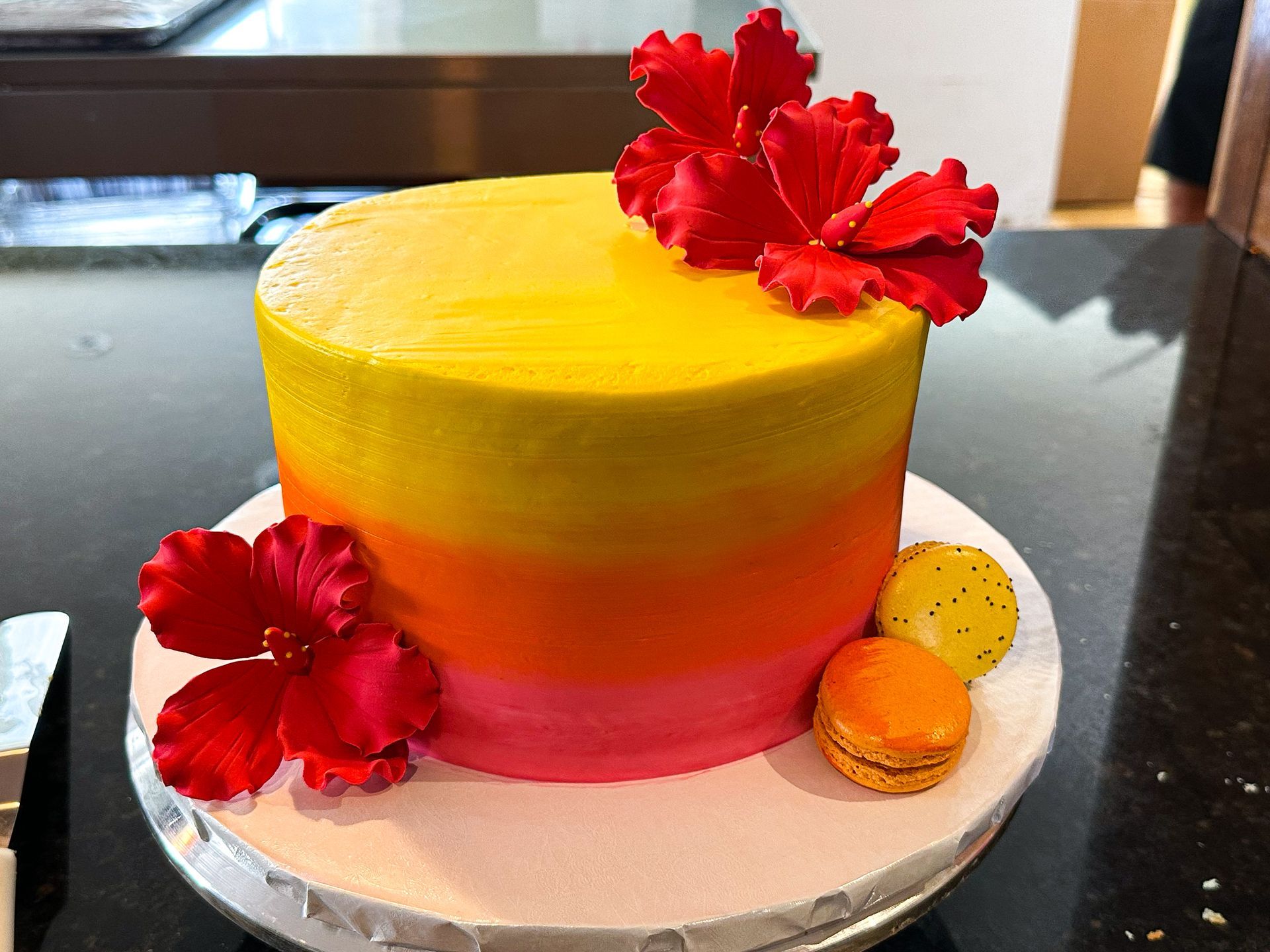 Cake with yellow, orange, and pink frosting, decorated with red hibiscus flowers and macarons.