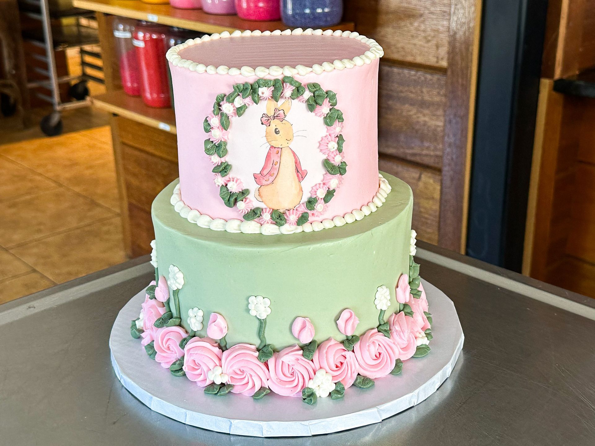 Two-tiered cake: pink top, green bottom, floral decorations, bunny illustration, on a metal surface.