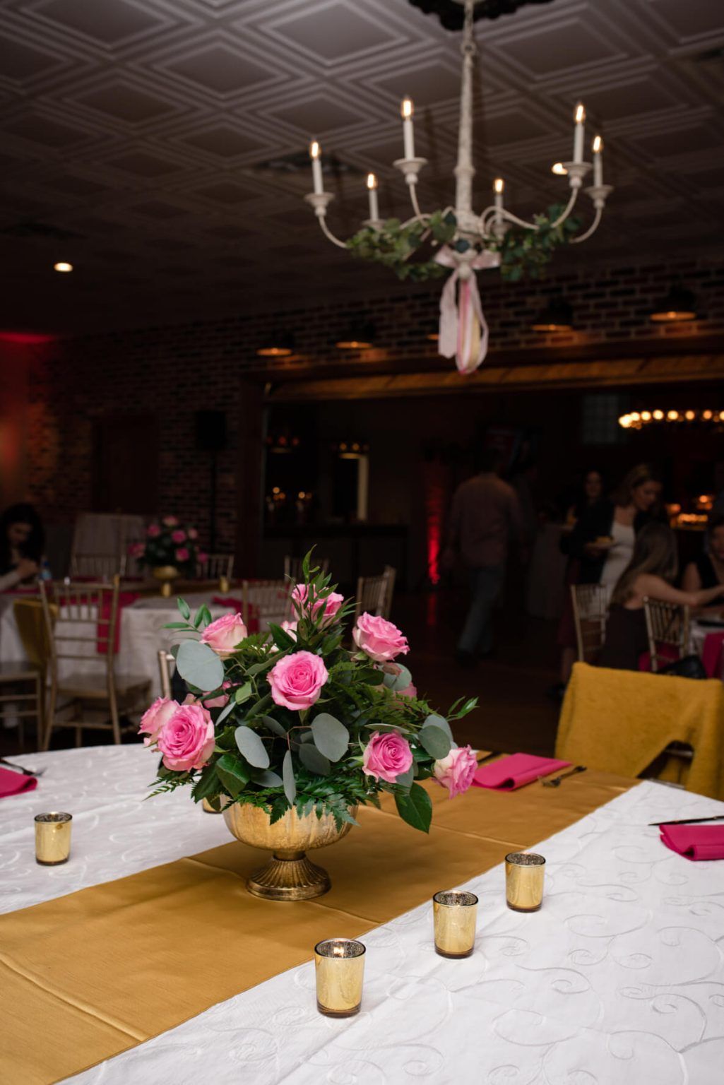 Table setting at event with pink roses in gold vase, gold runner, candles, and chandelier.
