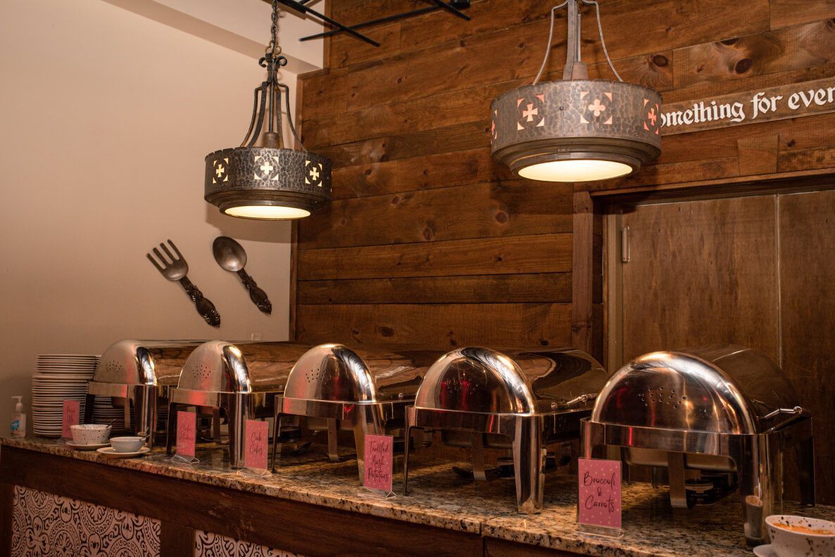Buffet serving station with stainless steel chafing dishes, wood paneling, and decorative lighting.