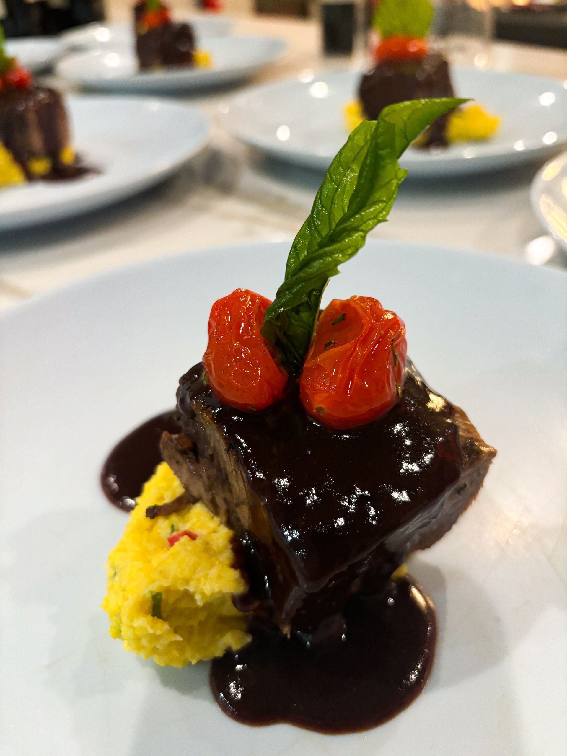 Gourmet dish: Braised meat cube with sauce, yellow puree, cherry tomatoes, and mint sprig on a white plate.