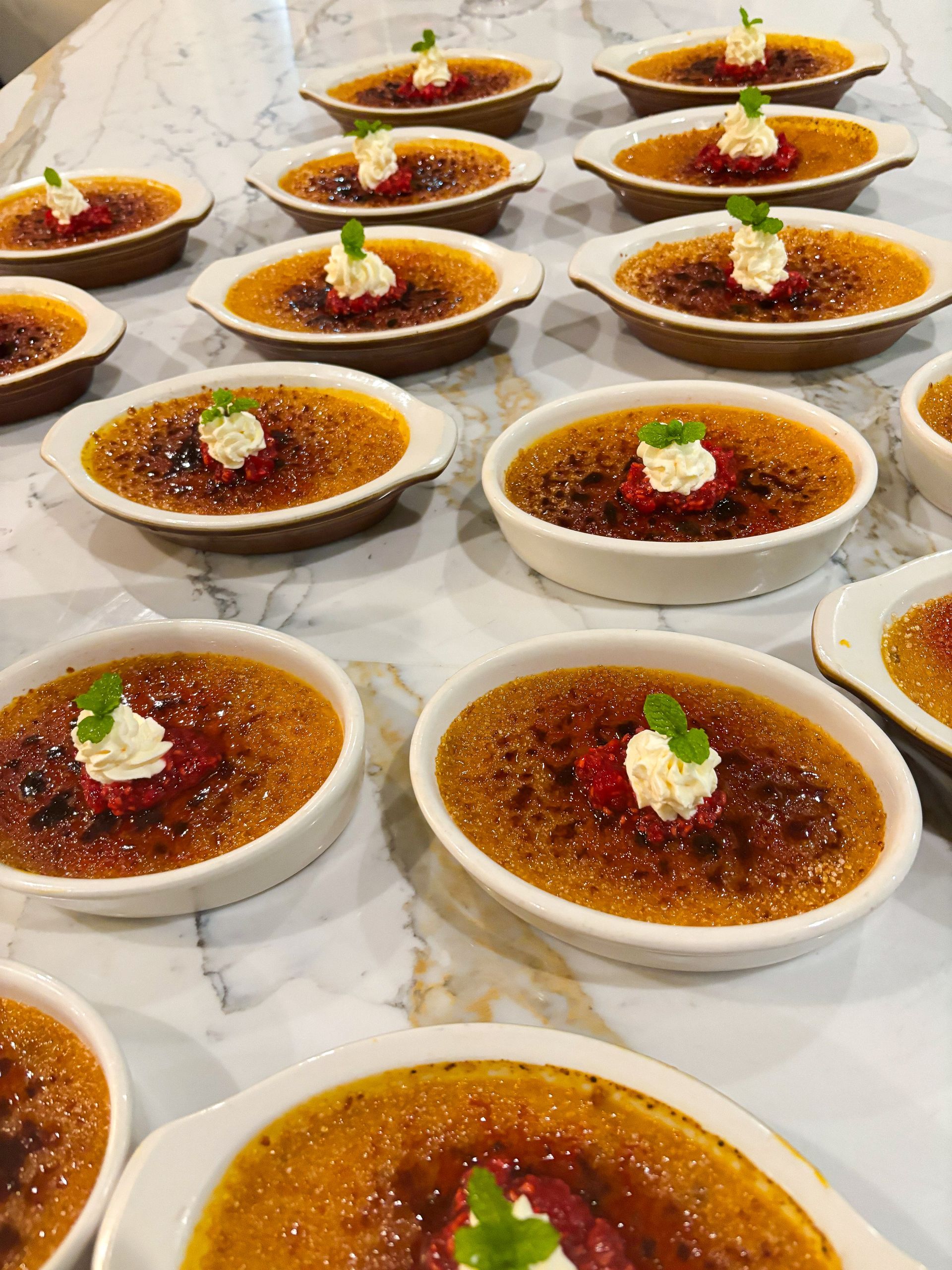 Creme brûlée desserts in oval ramekins, topped with whipped cream, raspberries, and mint, arranged on marble.
