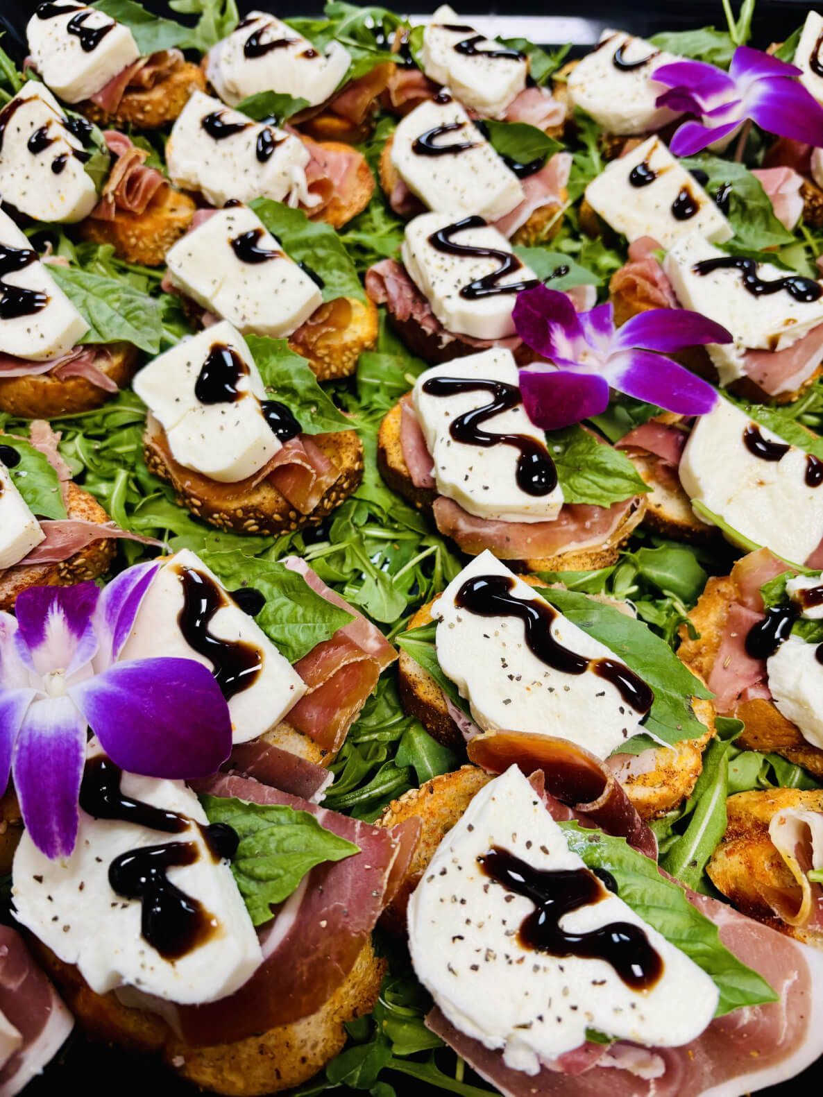 Tray of appetizers: bruschetta with prosciutto, mozzarella, basil, balsamic glaze, and orchid garnish.