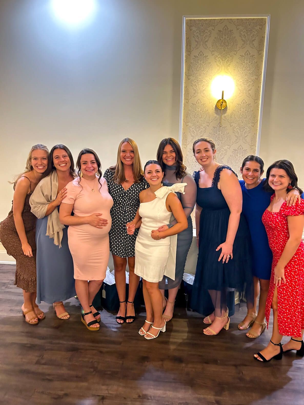 Group of women smiling together in a brightly lit room, wearing dresses.