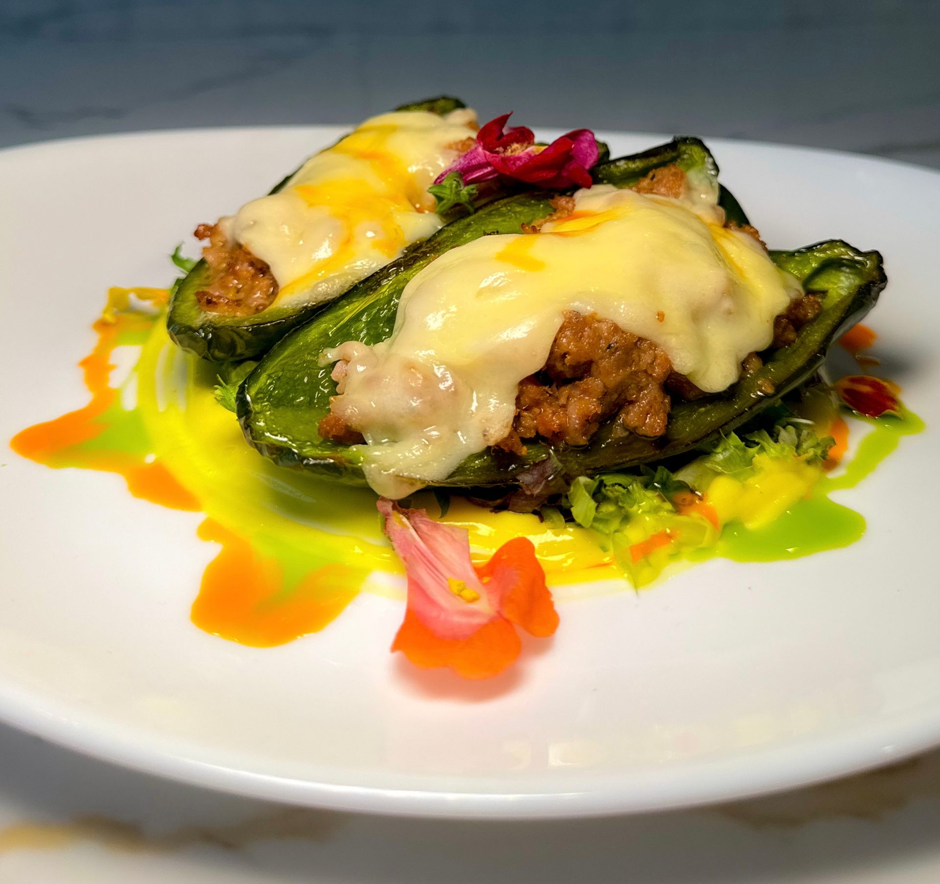 Stuffed poblano peppers topped with melted cheese, drizzled with colorful sauces, and garnished with flowers on a white plate.