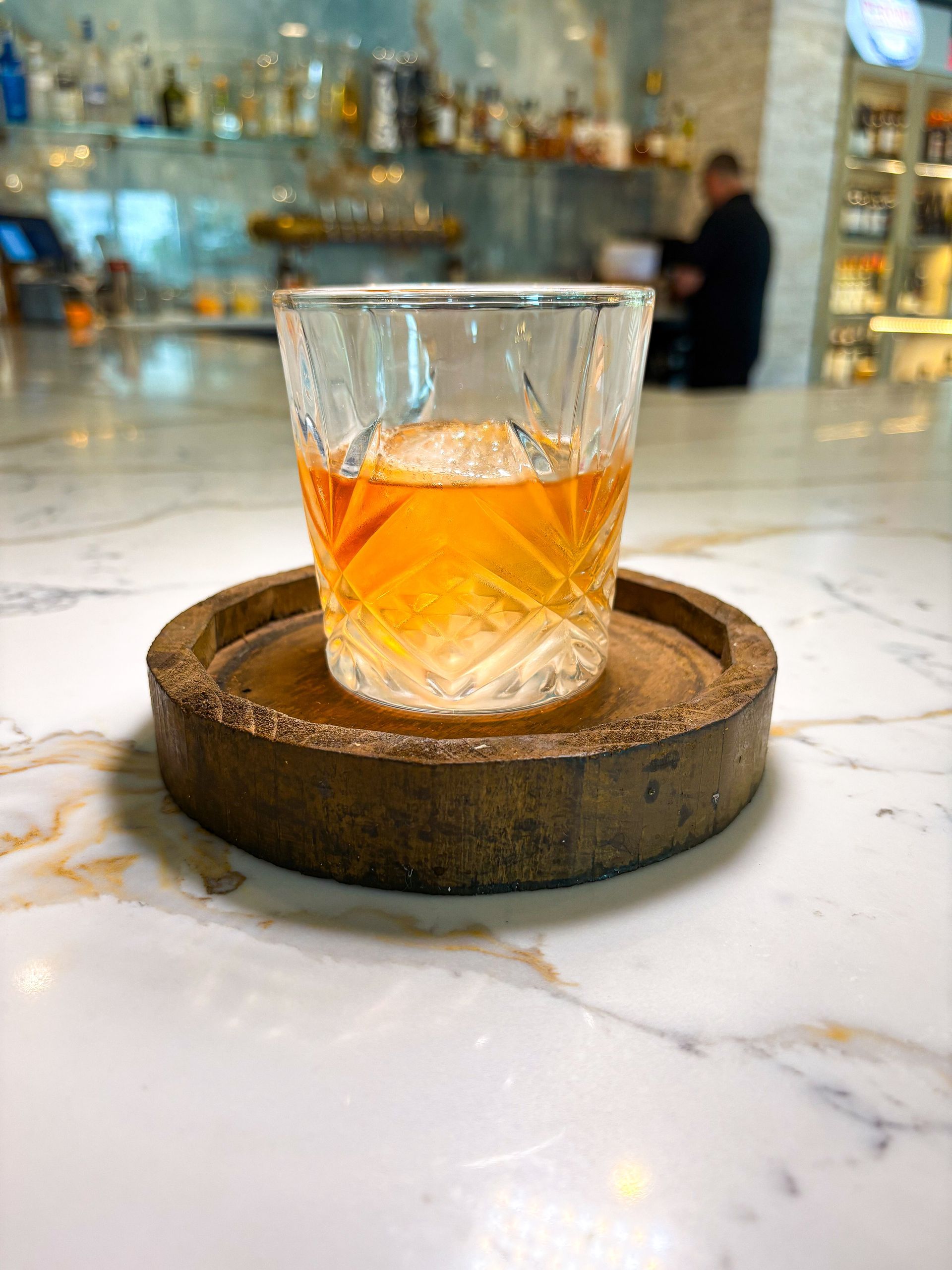 Cocktail on wooden coaster, marble bar, bar in background with bartender.