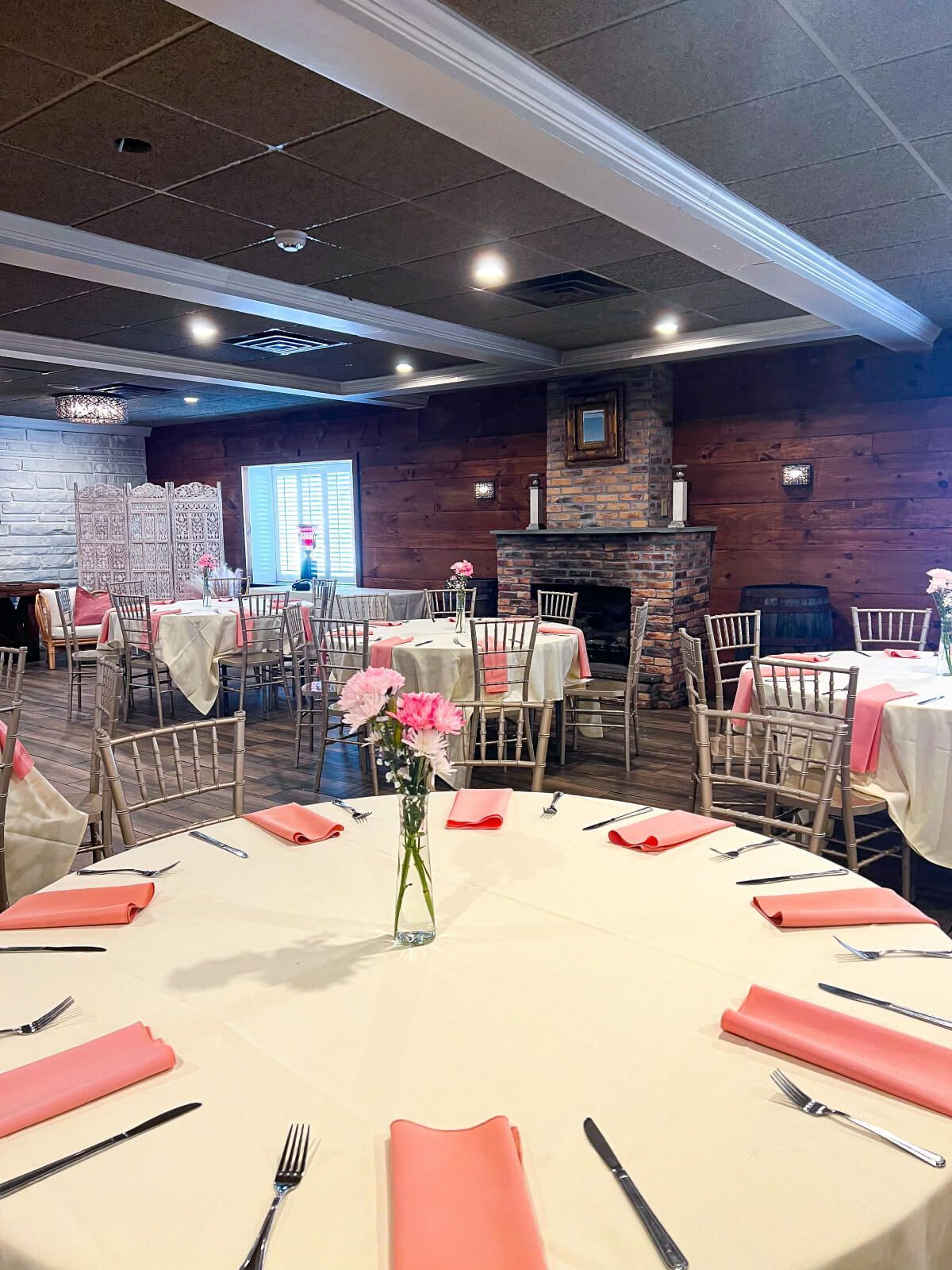 Round tables set for a gathering, with pink napkins and floral centerpieces, in a room with a fireplace.