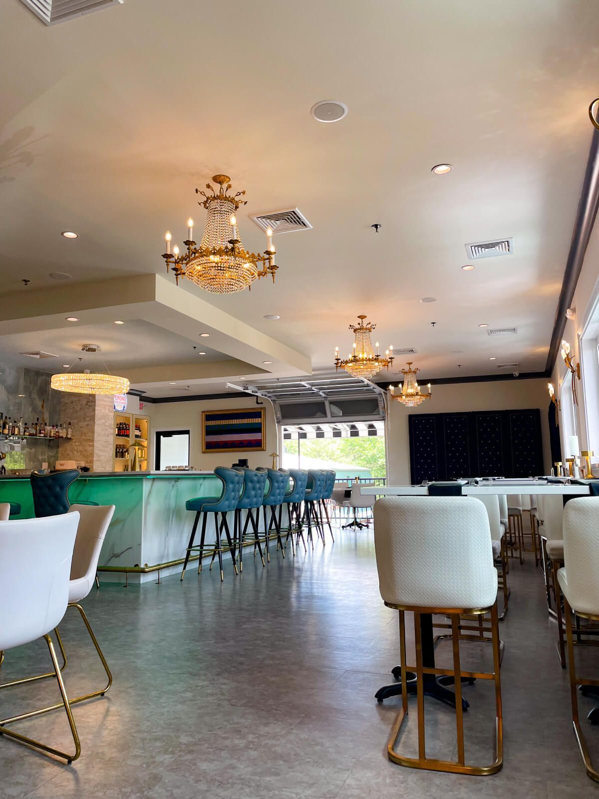 Interior of a bar/restaurant with chandelier lights, teal bar, and white tables and chairs.