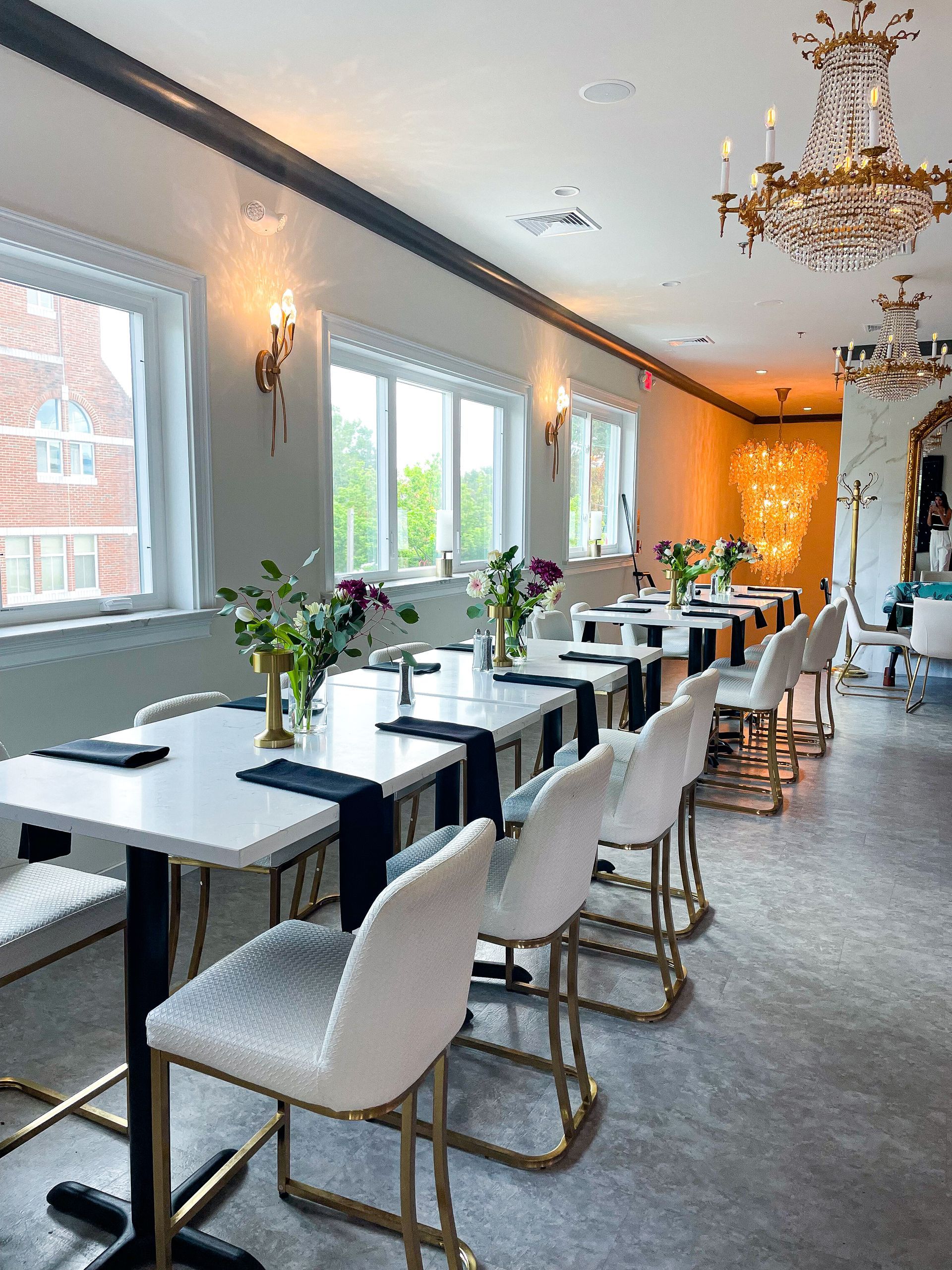 Restaurant interior with white tables, chairs, and chandeliers. Windows line the wall.