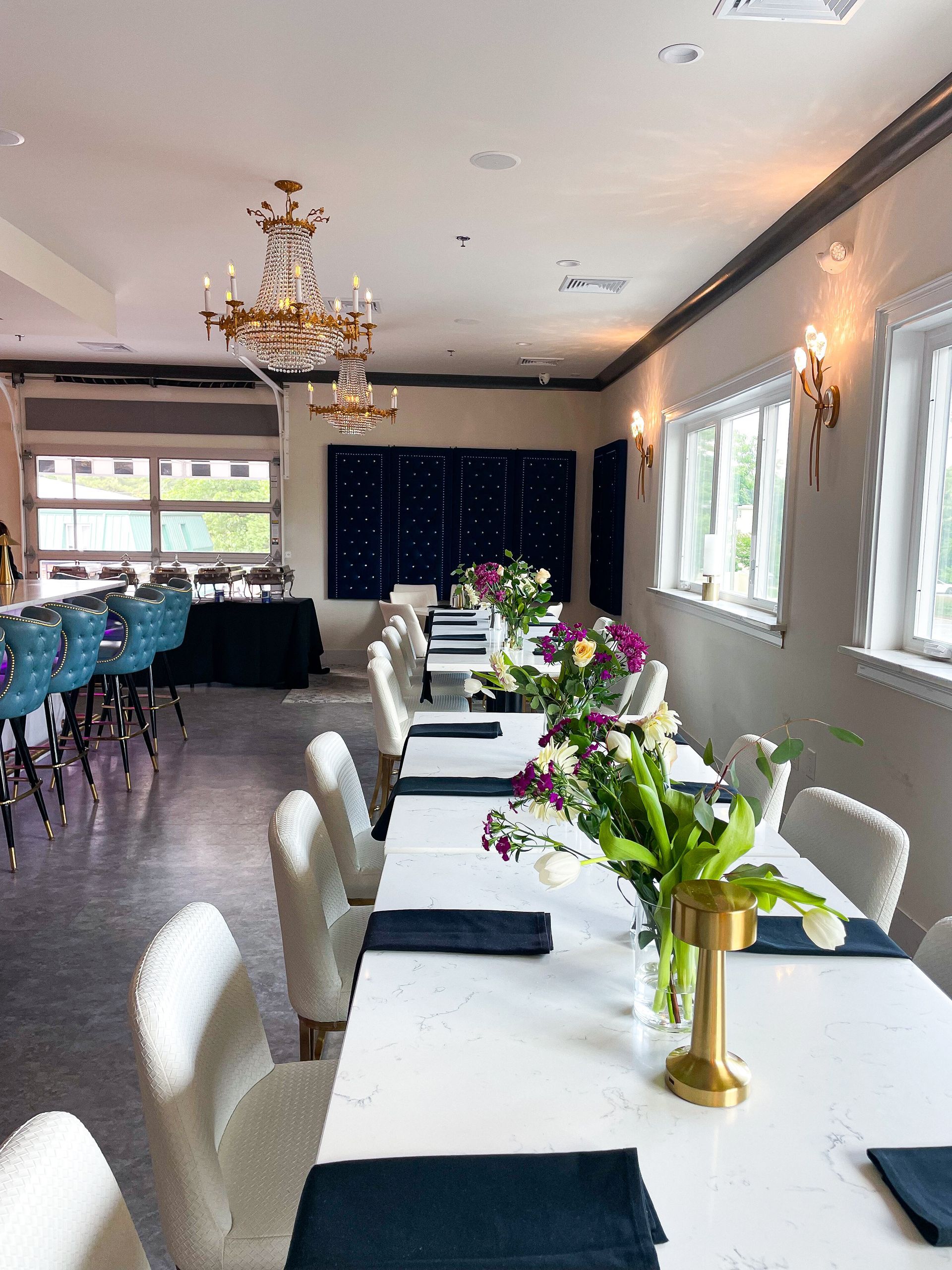 Long dining tables set for a banquet with white chairs, floral centerpieces, and a bar area.