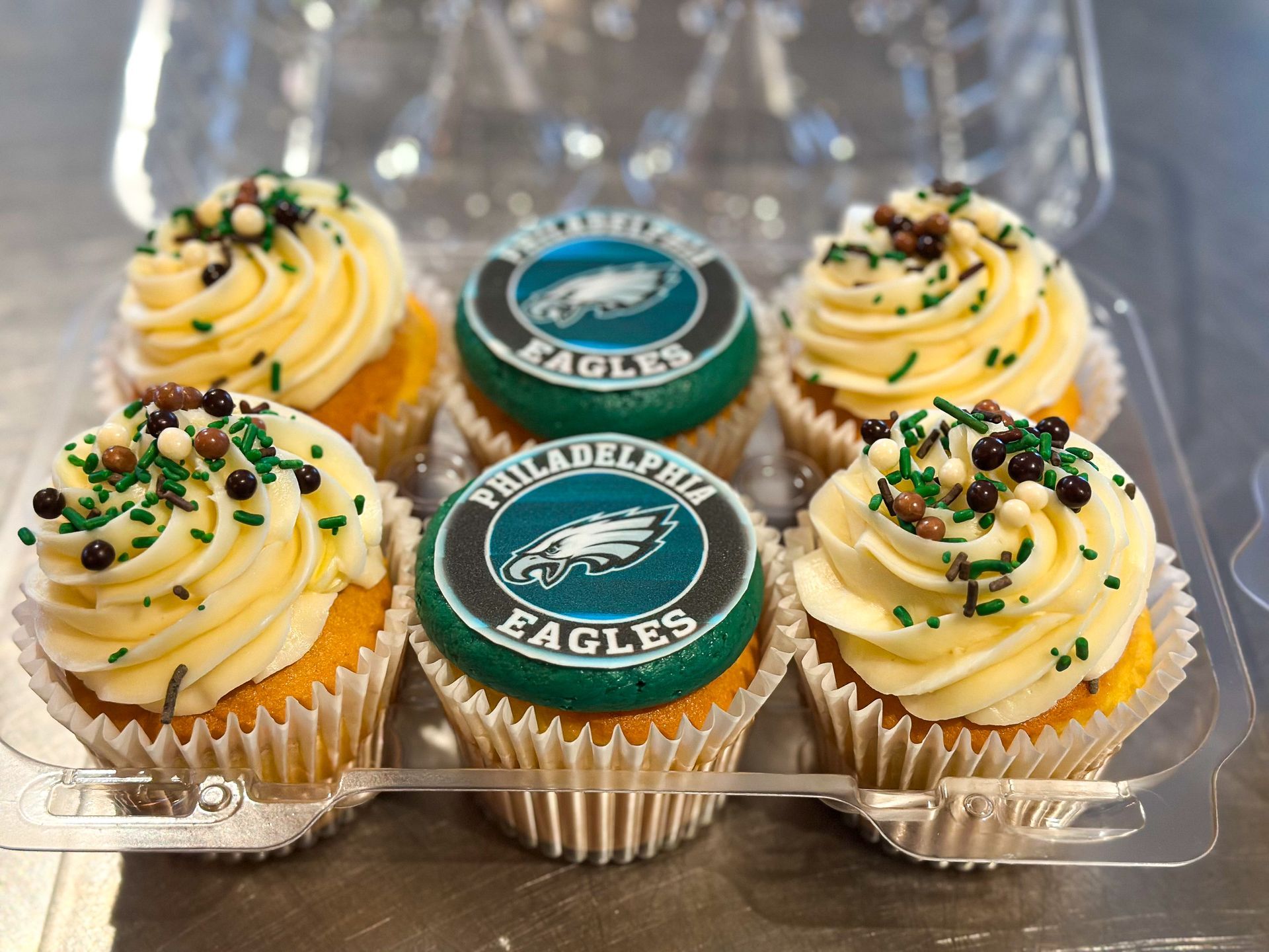Six Philadelphia Eagles cupcakes in a plastic container. Yellow frosting, team logo toppers, and sprinkles.