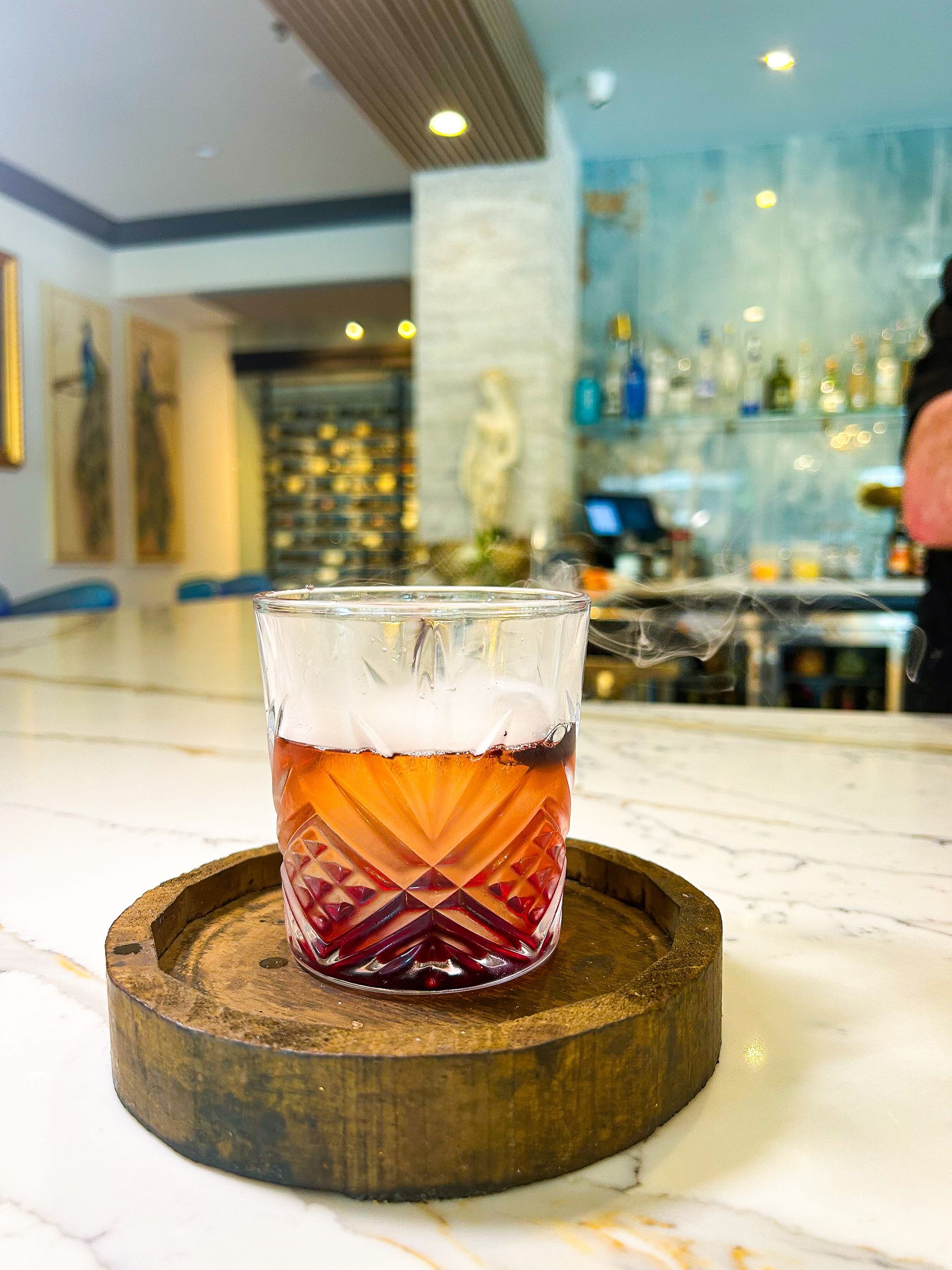 Cocktail in a patterned glass on a wooden coaster at a bar, with bottles and a person in the background.
