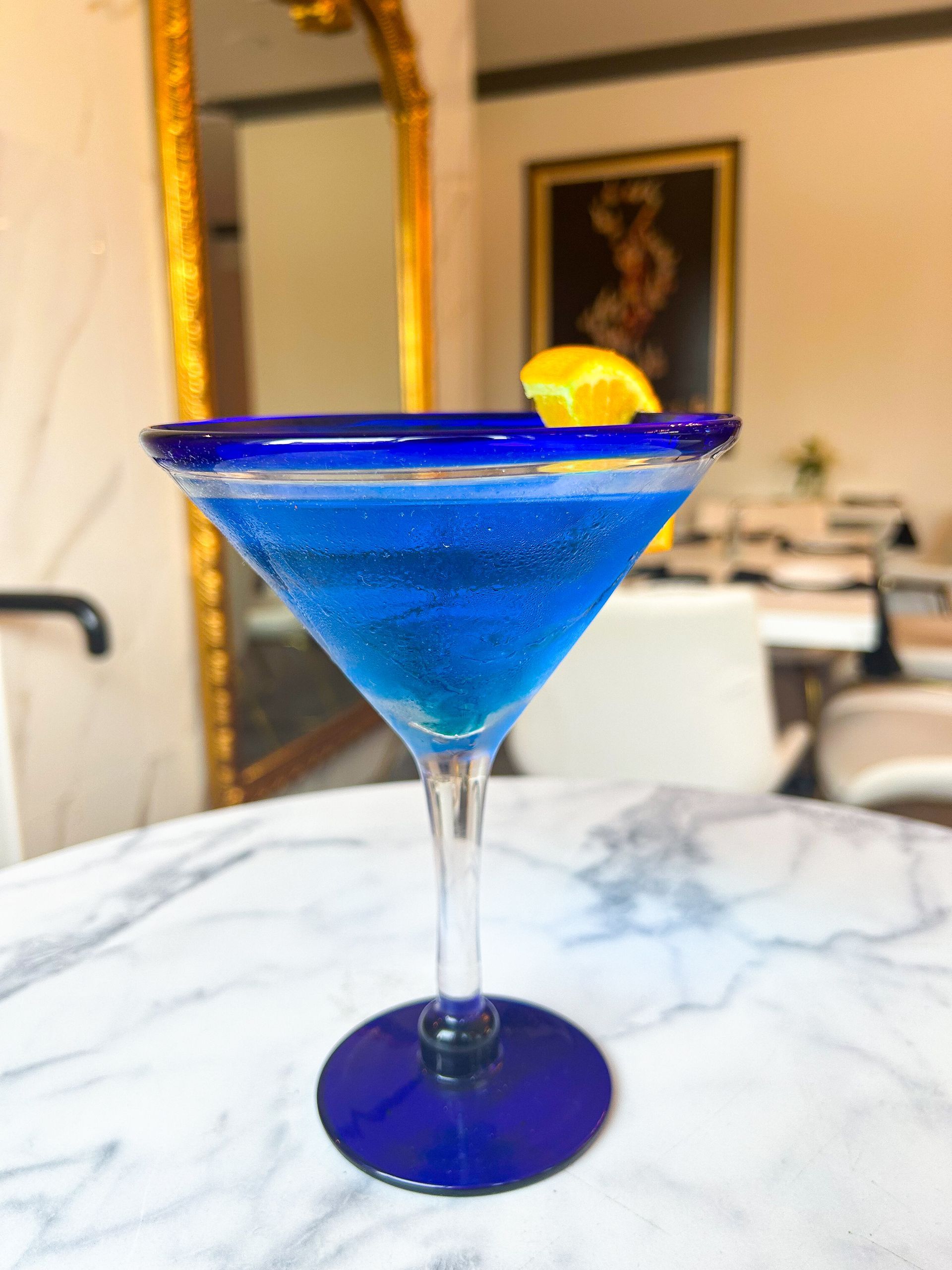 Blue cocktail in a martini glass with a lemon slice garnish, on a marble table.