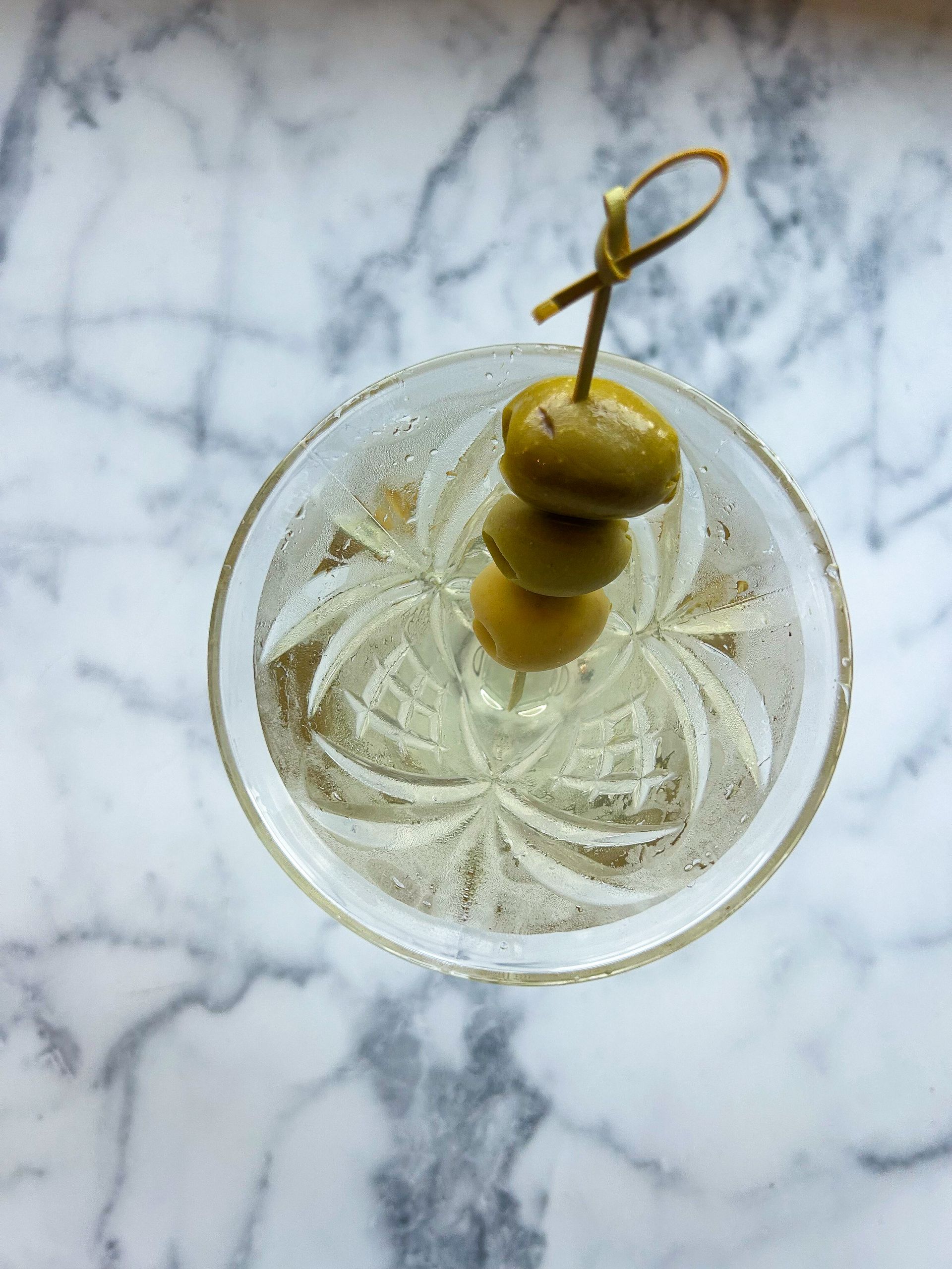 Martini in a crystal glass garnished with three olives on a skewer, on a marble surface.
