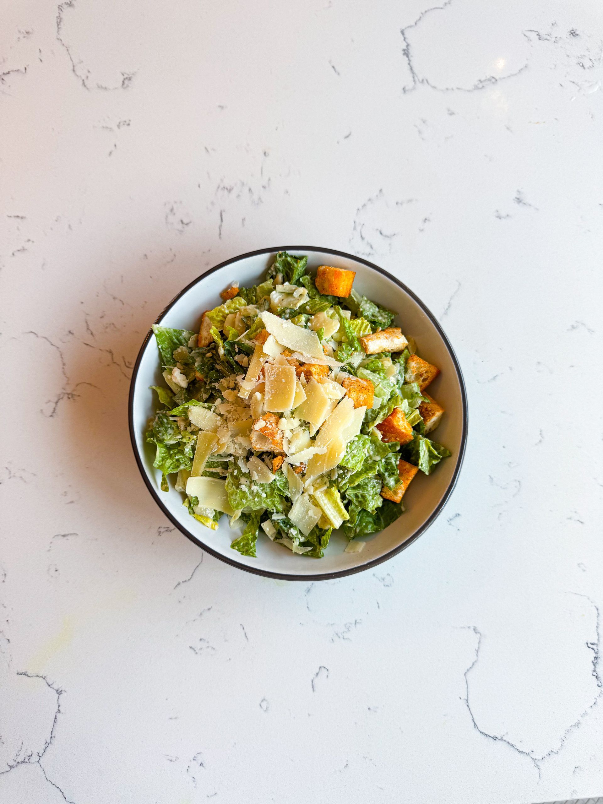 Caesar salad in a white bowl, with parmesan cheese, croutons, and romaine lettuce on a white countertop.
