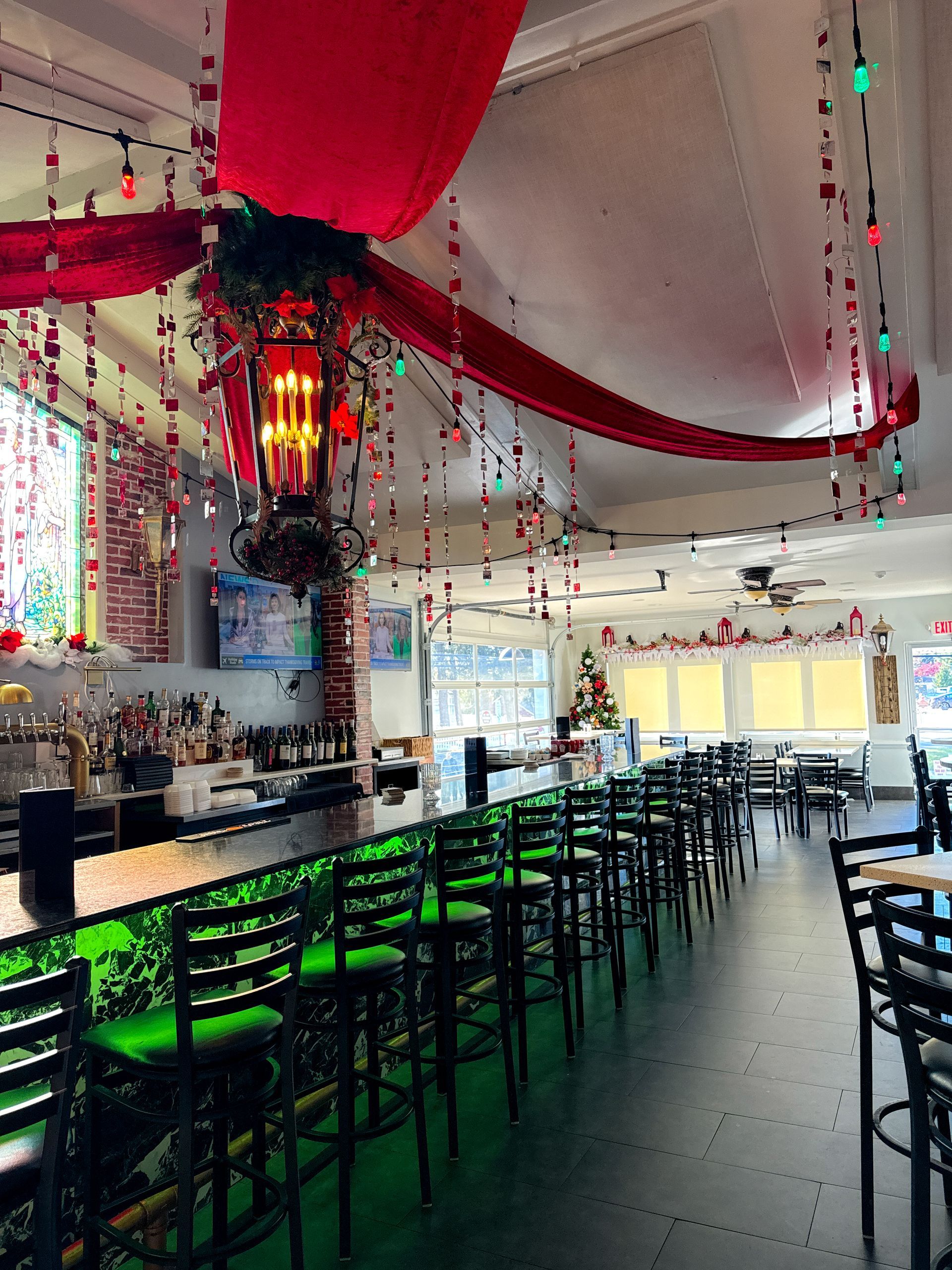 Interior of a restaurant decorated for a holiday. Red ribbons and lights hang from the ceiling. A green-lit bar and chairs are in view.
